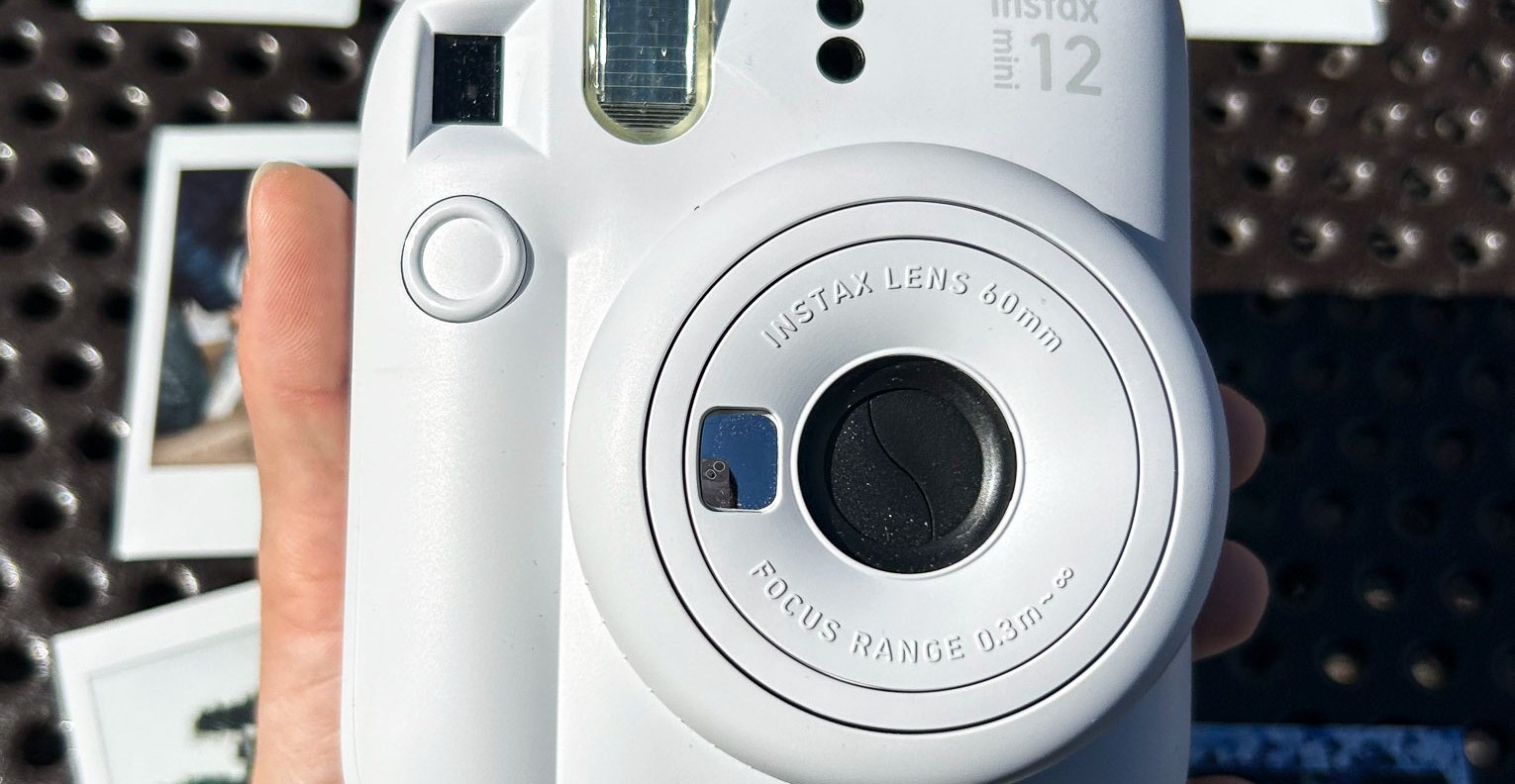 A closeup of Fujifilm’s Instax Mini 12 camera and its lens structure surrounded by photos it printed in the background.