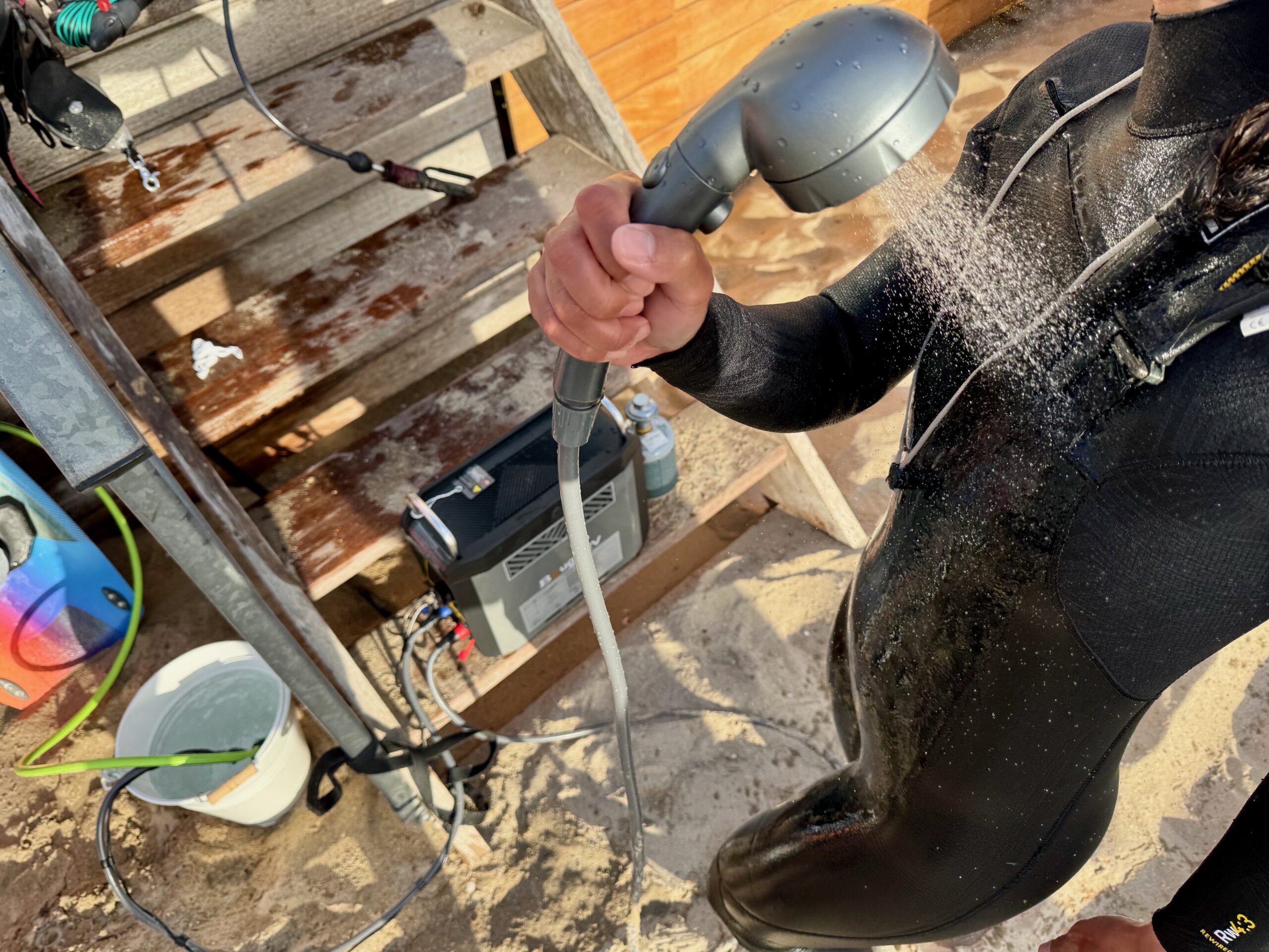 A person wearing a wetsuit at the beach, standing in front of the BougeRV water heater sitting on some stairs, holds the shower head in her hand and directs the spray at her chest.