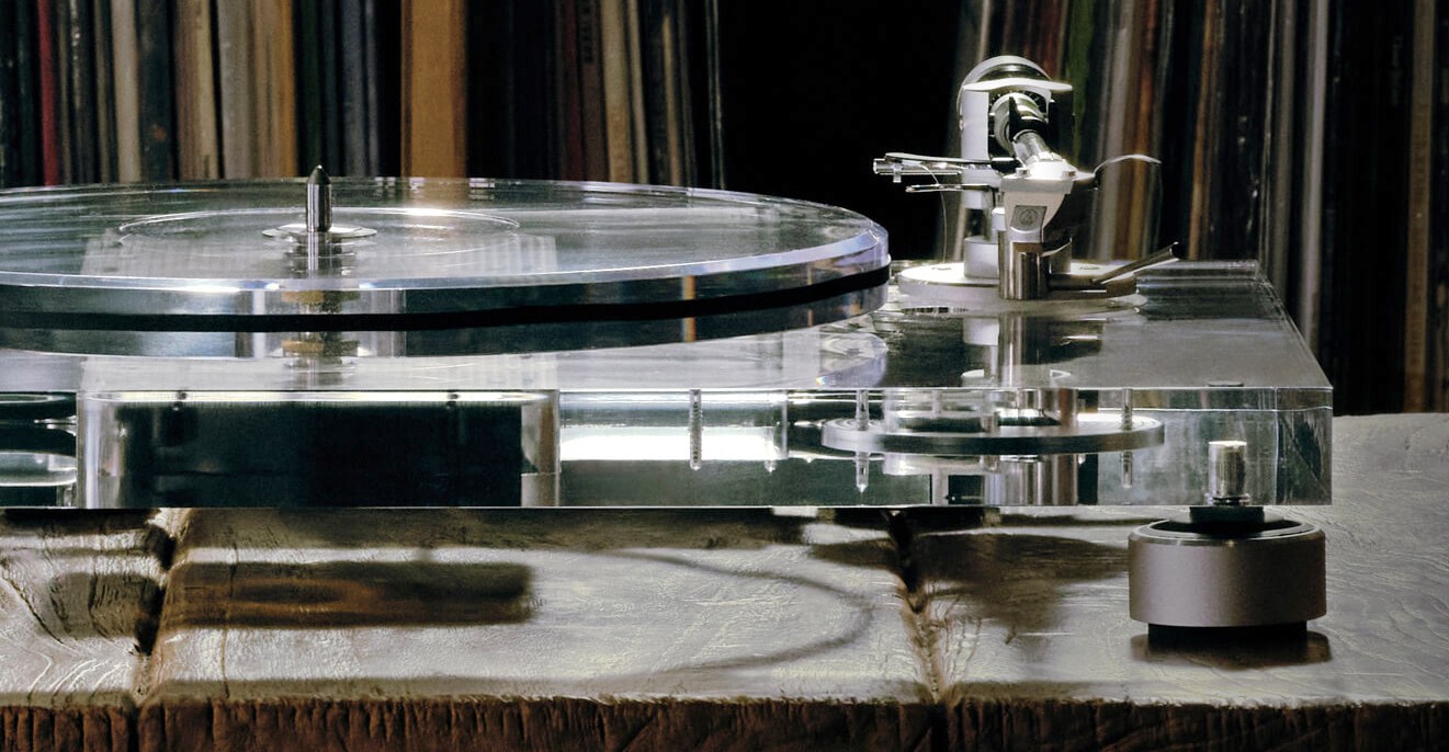 Audio-Technica’s transparent turntable on a wooden stand in front of a shelf full of record sleeves.
