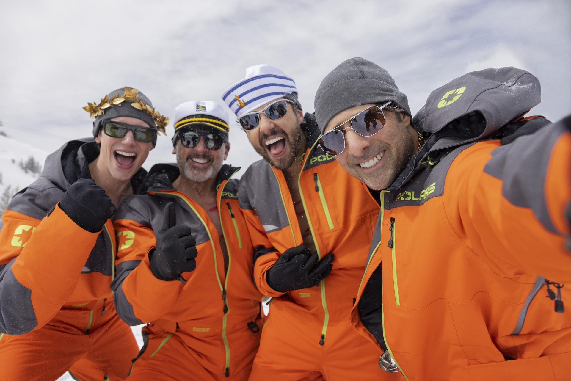 Four men in matching orange snowsuits smiling while taking a photo together on a mountain.
