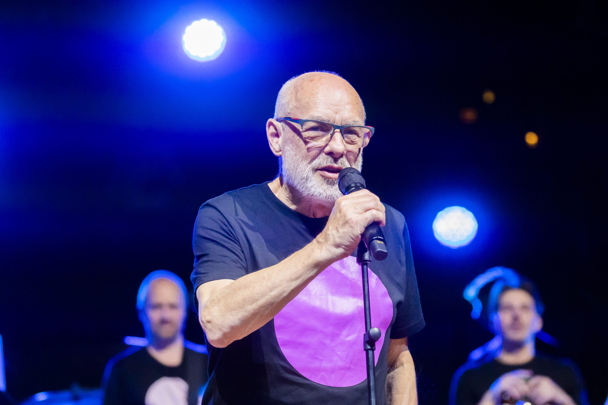 Brian Eno holds a microphone at a mic stand