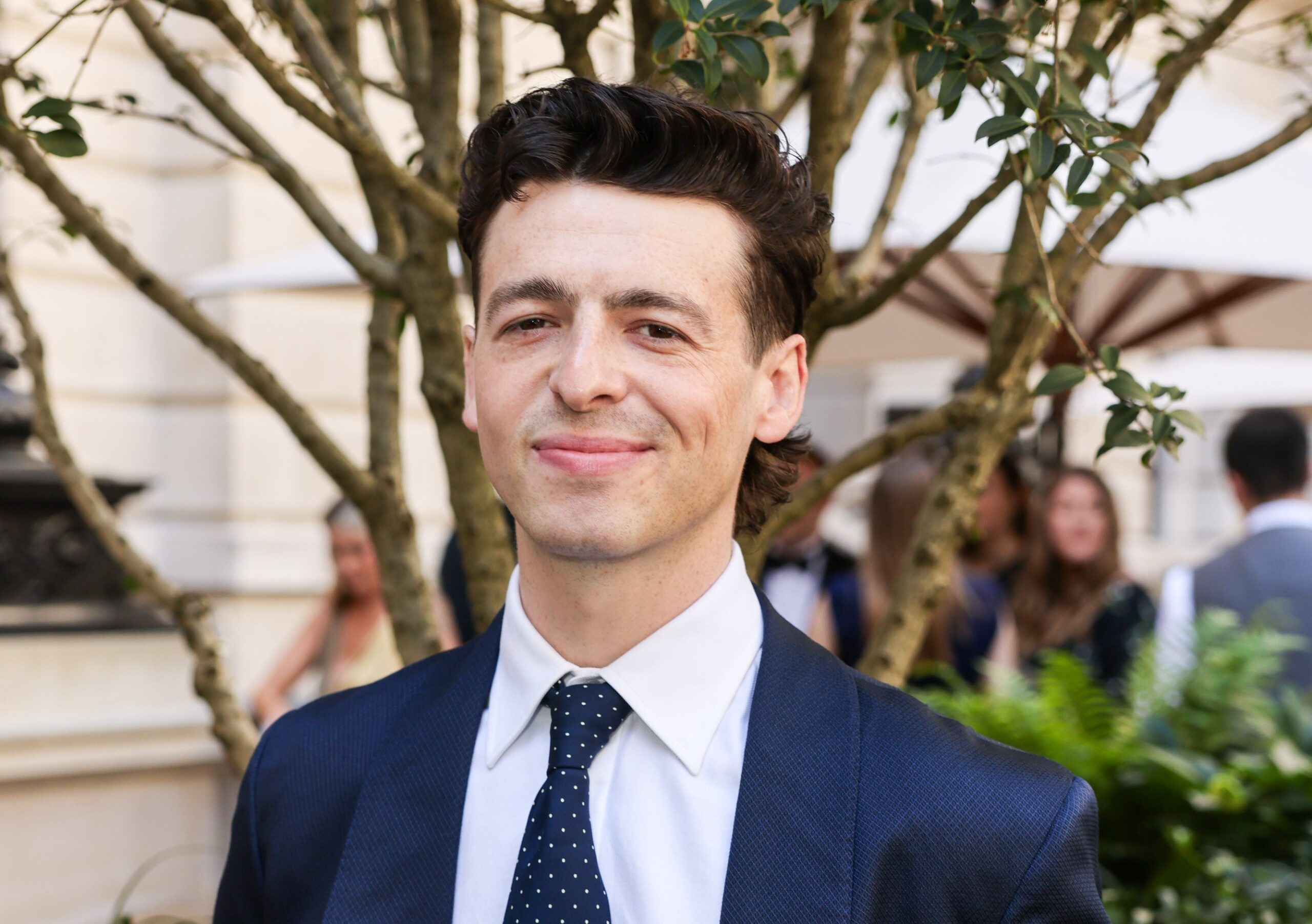 A man in a blue suit and tie standing in front of a tree and some bushes.