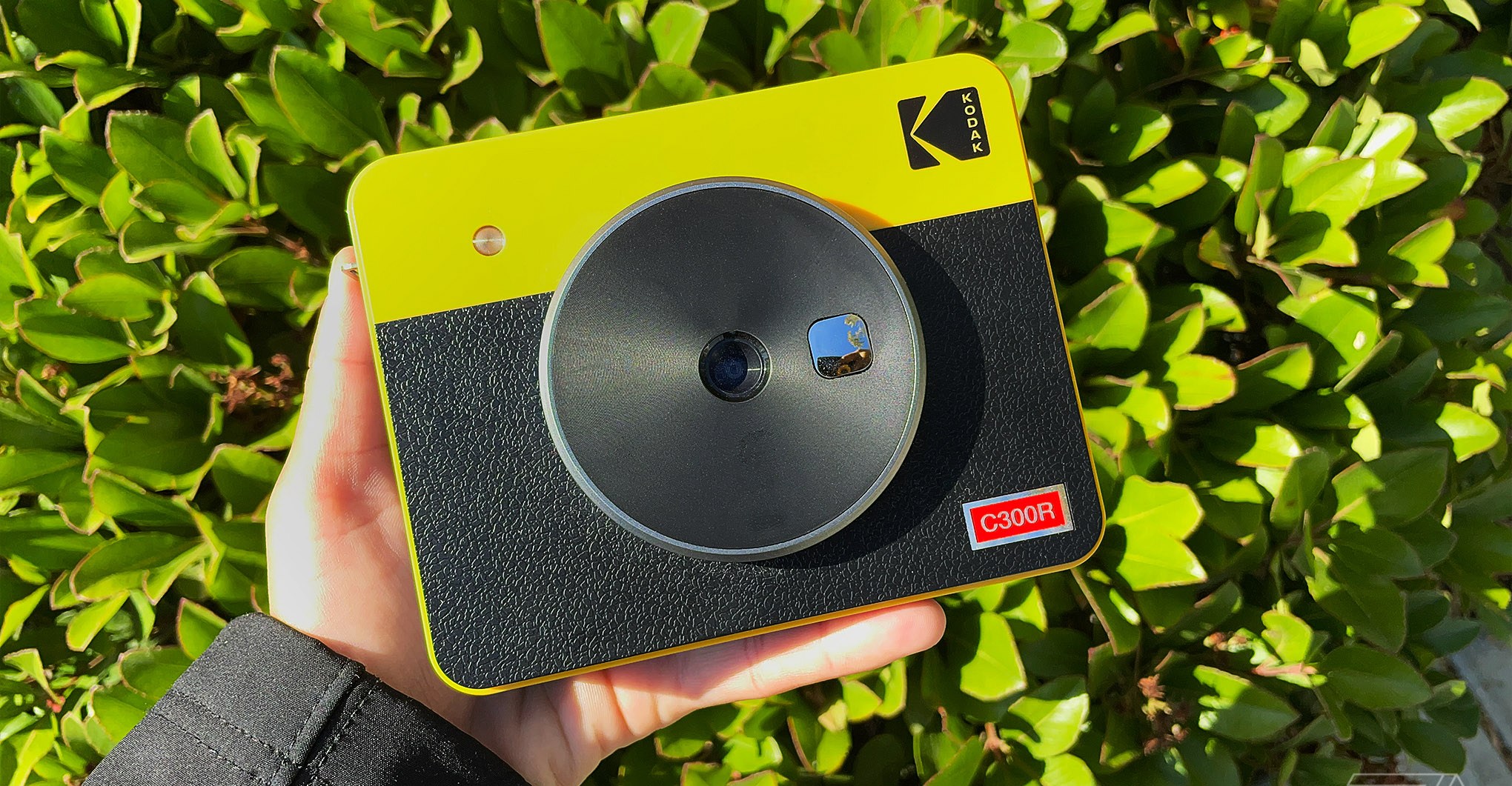 The yellow and black Kodak Mini Shot 3 Retro being held up sideways with a hand and a bush as the background.