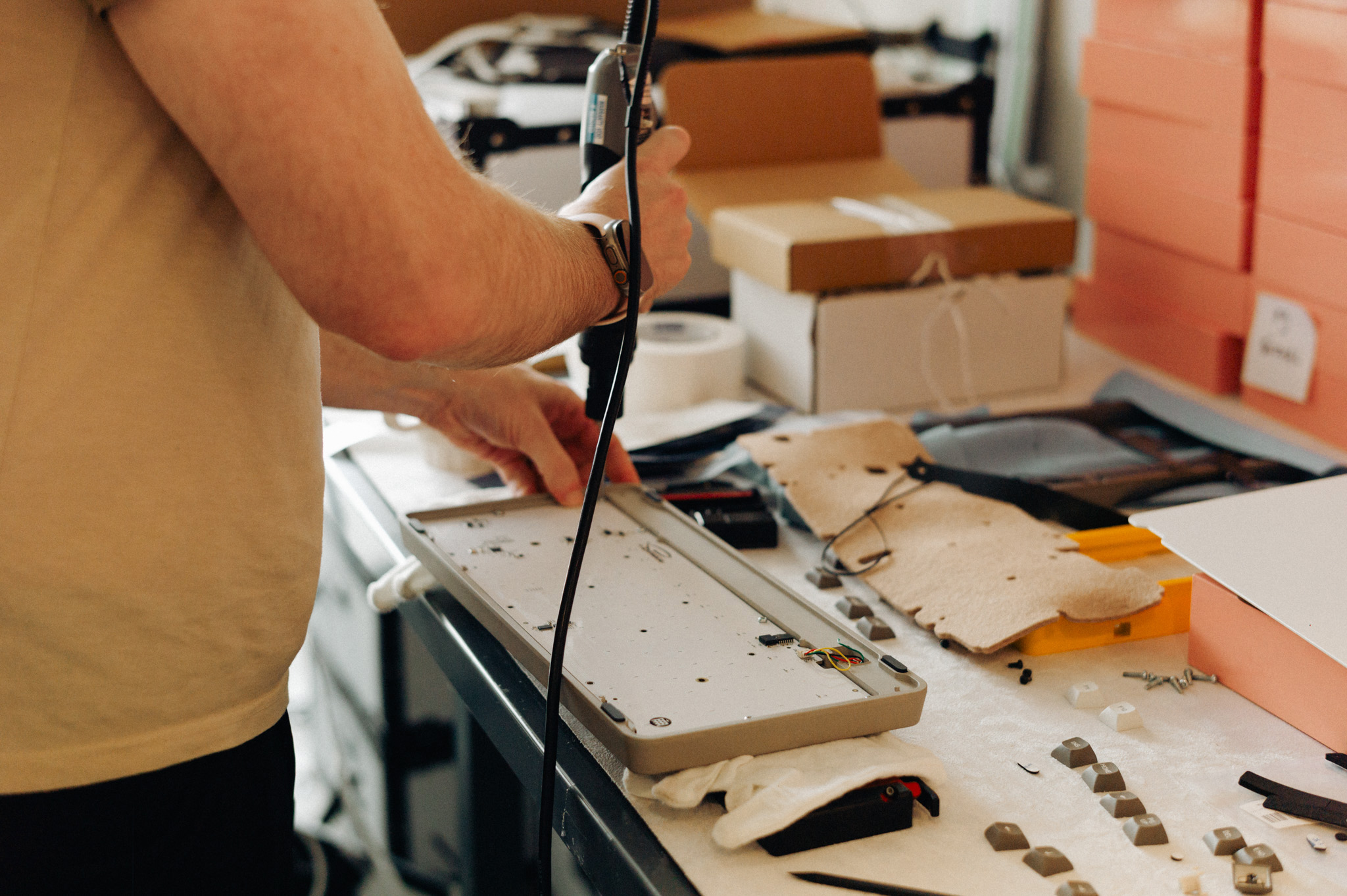 side view of a white man in a cluttered workshop using an electric screwdriver to assemble a keyboard. 