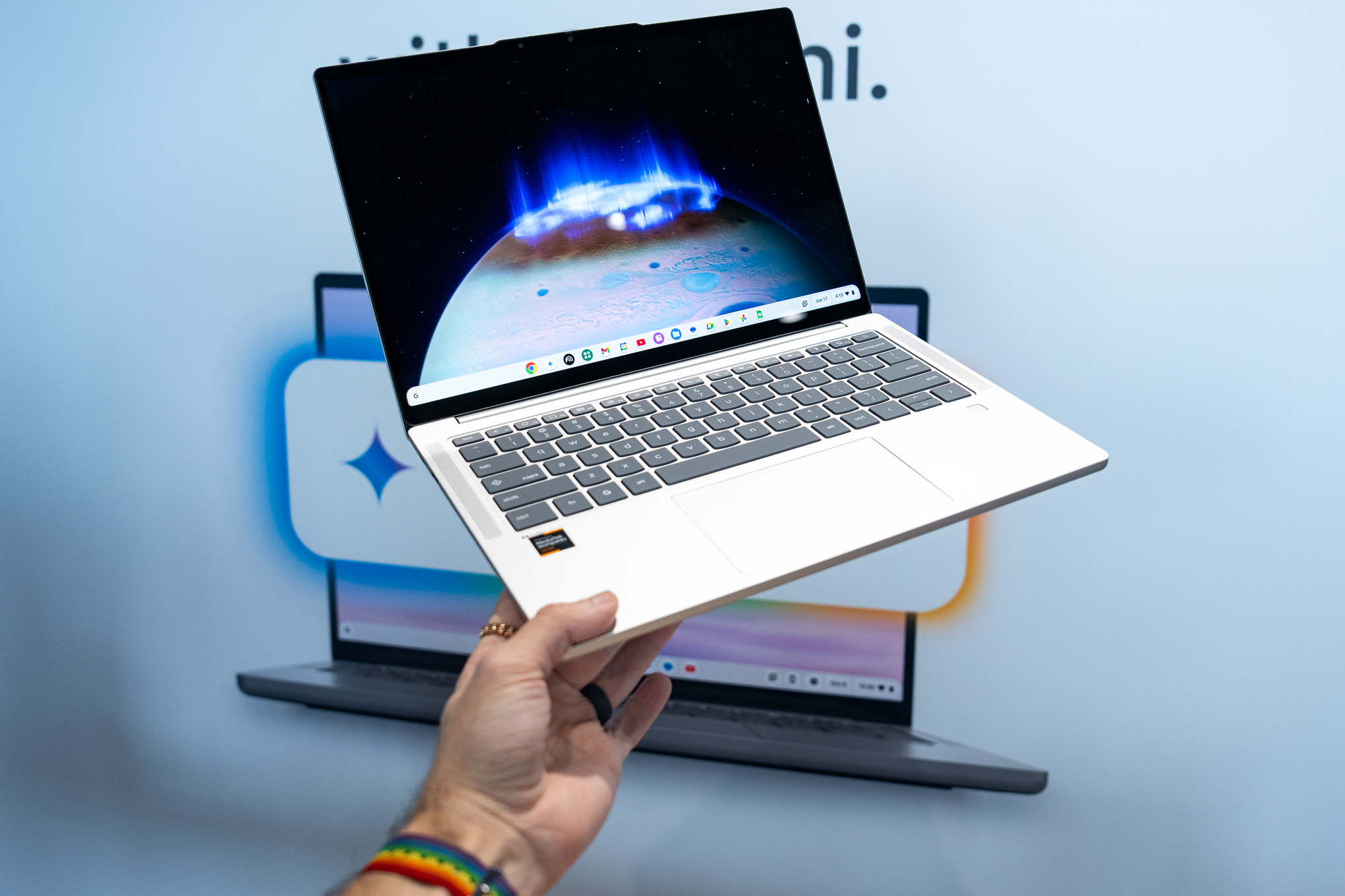 A hand holding a silver Lenovo Chromebook Plus 14 laptop up at an event, in front of a backdrop.