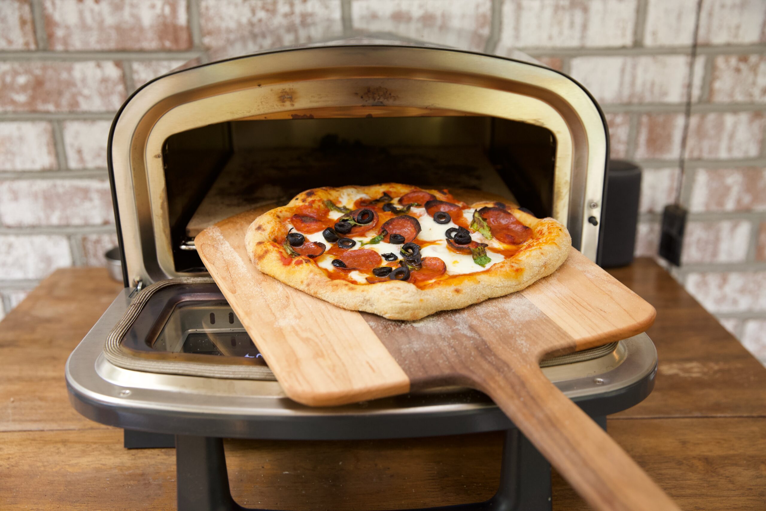 A pizza on a wooden pizza peel being removed from a pizza oven on a wooden table with a brick background.