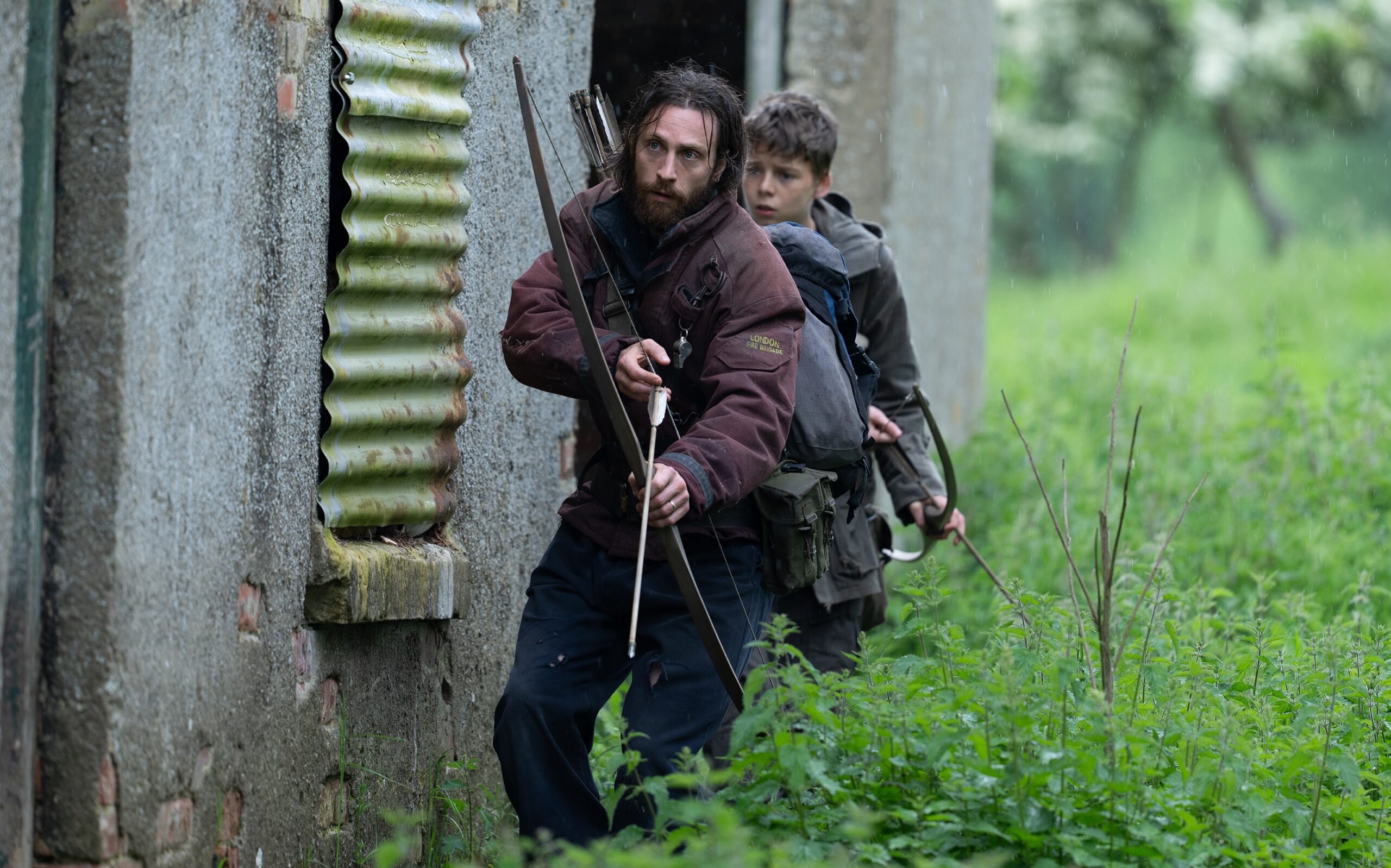 A man and a boy both holding longg bows as they sidle up against a building.