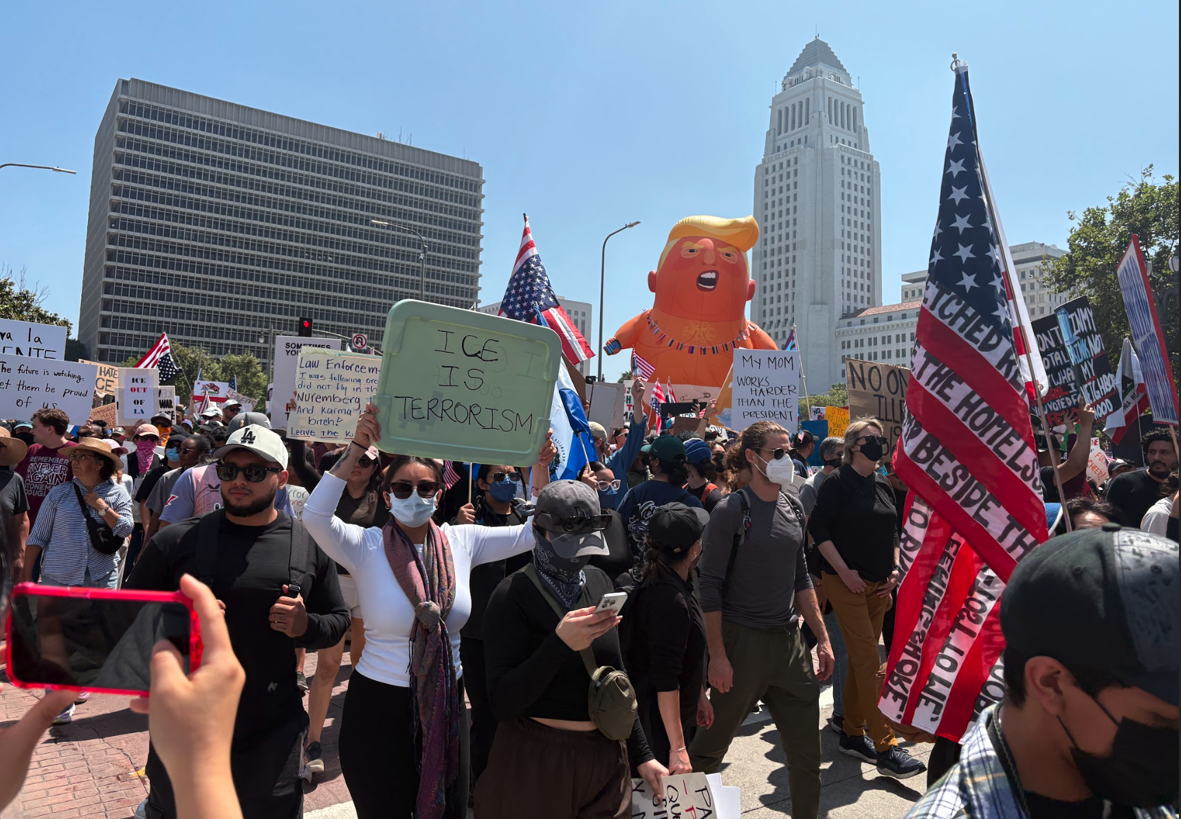 A parade balloon depicting Donald Trump as a baby wearing a diaper floats above a crowd of people in front of City Hall.￼