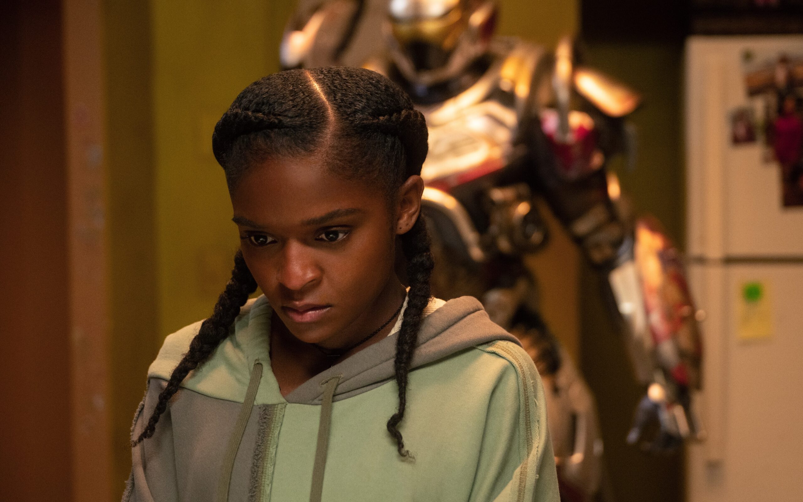 A girl wearing a gray and green hoodie. Behind the girl is a suit of high-tech armor.