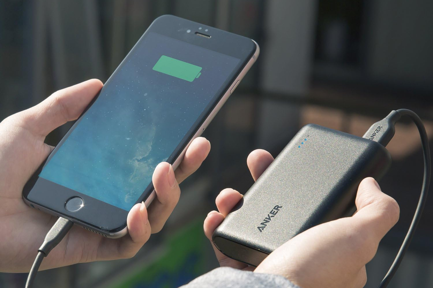 A hand holding a smartphone connected to an Anker power bank with a USB cable.