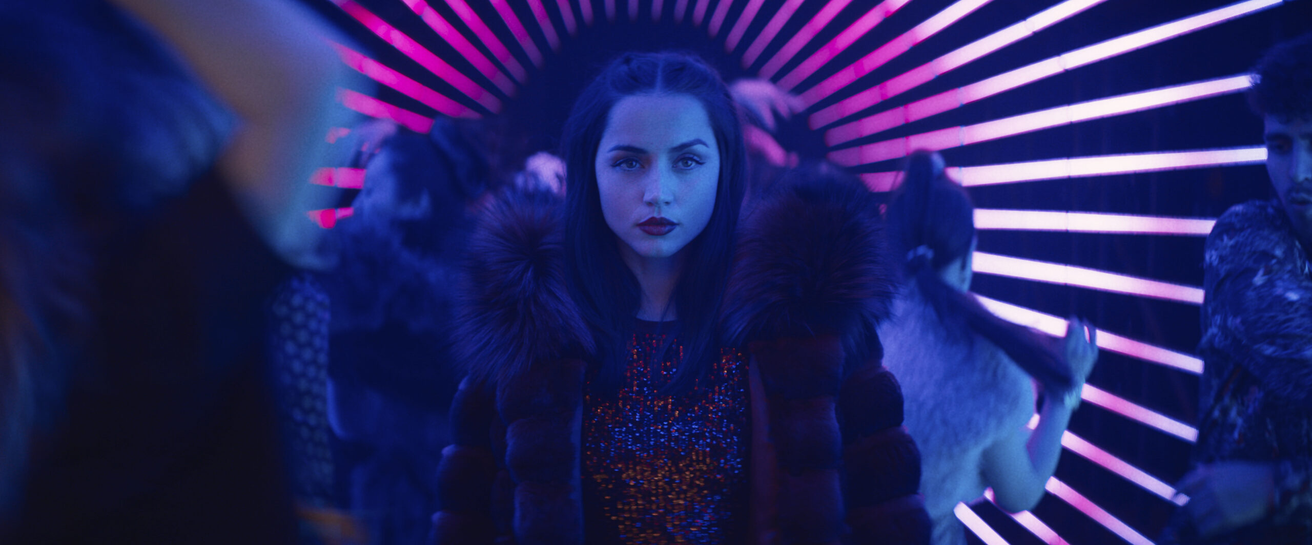 A woman wearing a fur coat and walking through a dark hallway illuminated by glowing pink light strips.