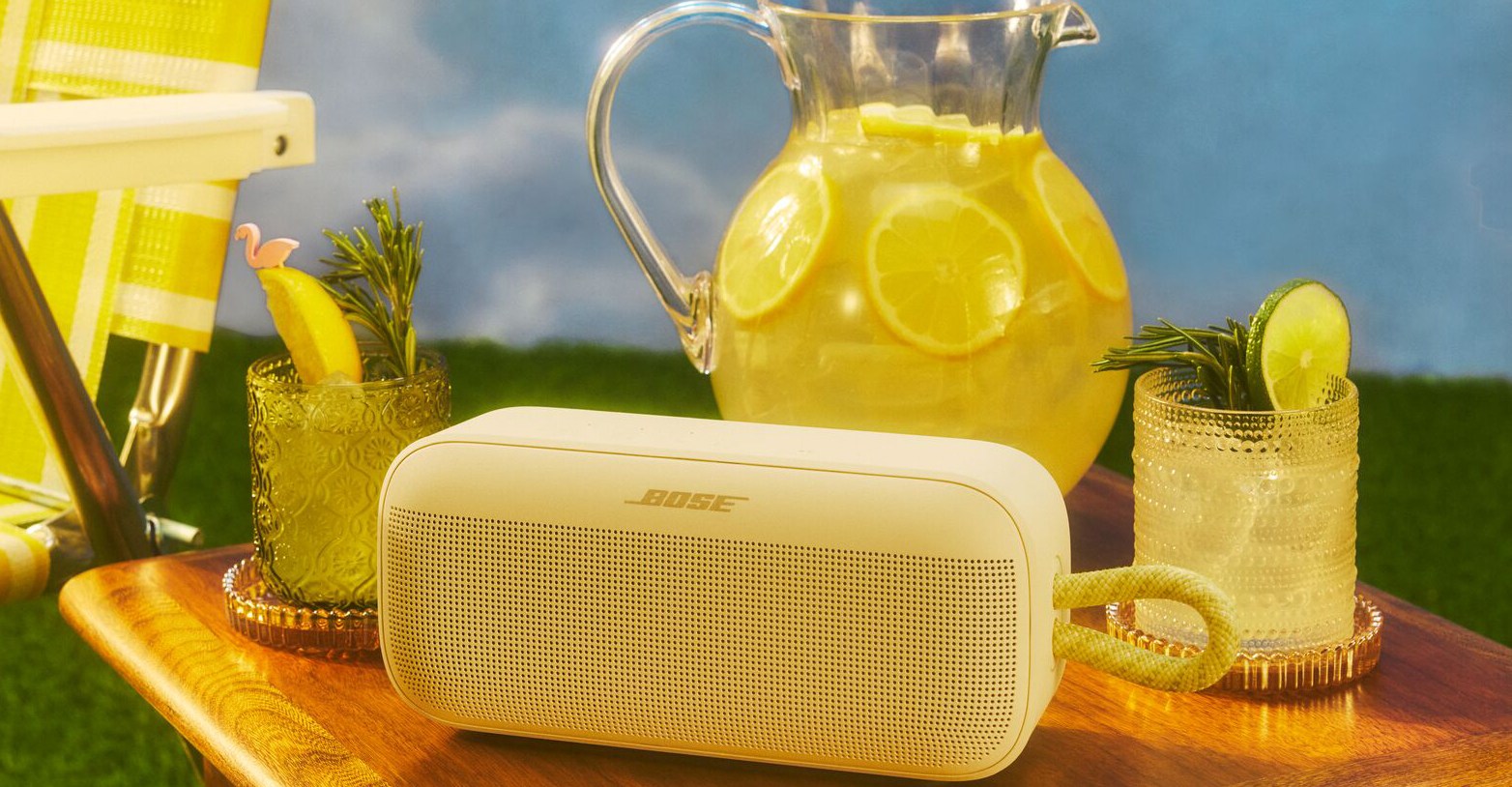 The Bose SoundLink Plus portable speaker on a wooden table surrounded by yellow objects including a pitcher of lemonade.