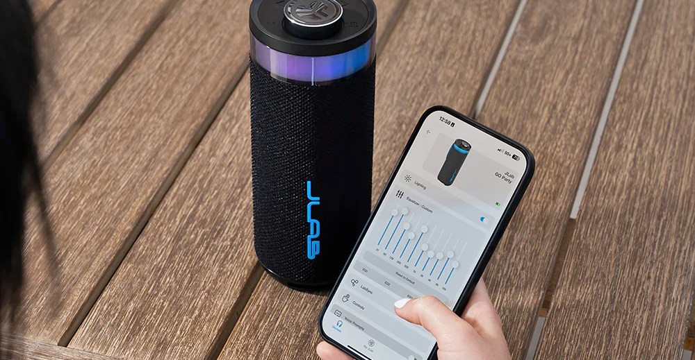 A person adjusts settings on a mobile app on a smartphone next to a JLab Bluetooth speaker sitting on a wood table.