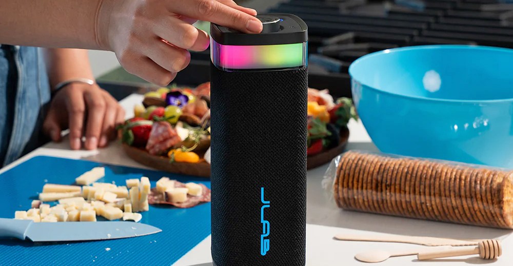 A person turns a knob on top of a JLab Bluetooth speaker sitting on a table full of food.