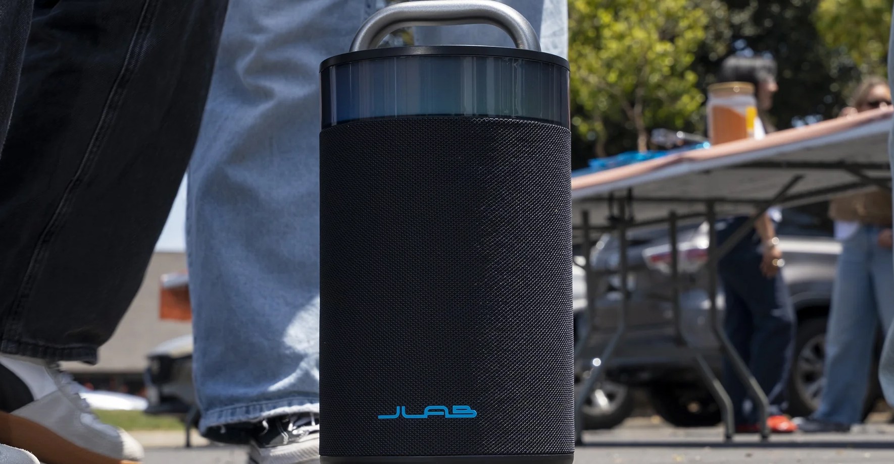 Several people standing around a JLab Bluetooth speaker sitting on the ground.
