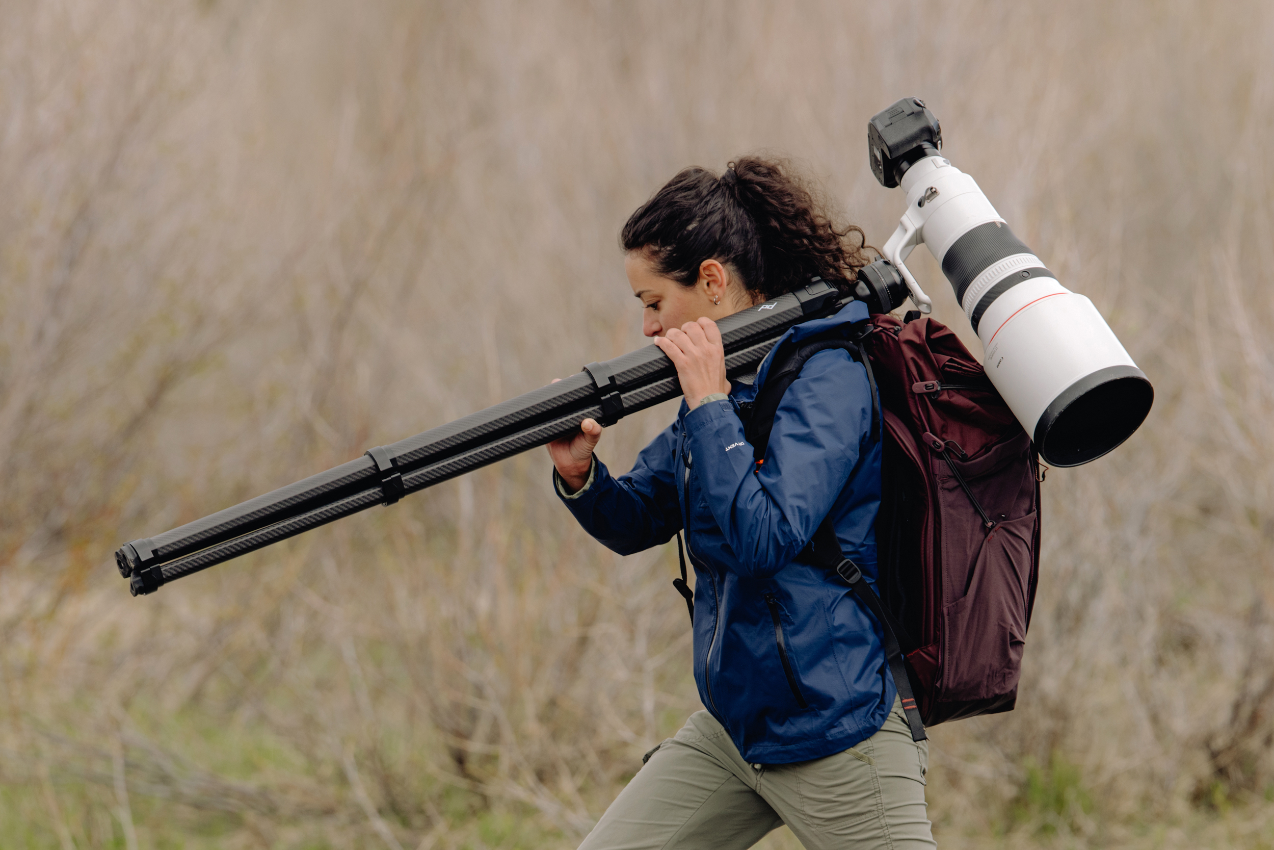 A photographer carrying a camera with a large lens attached to a Peak Design tripod.