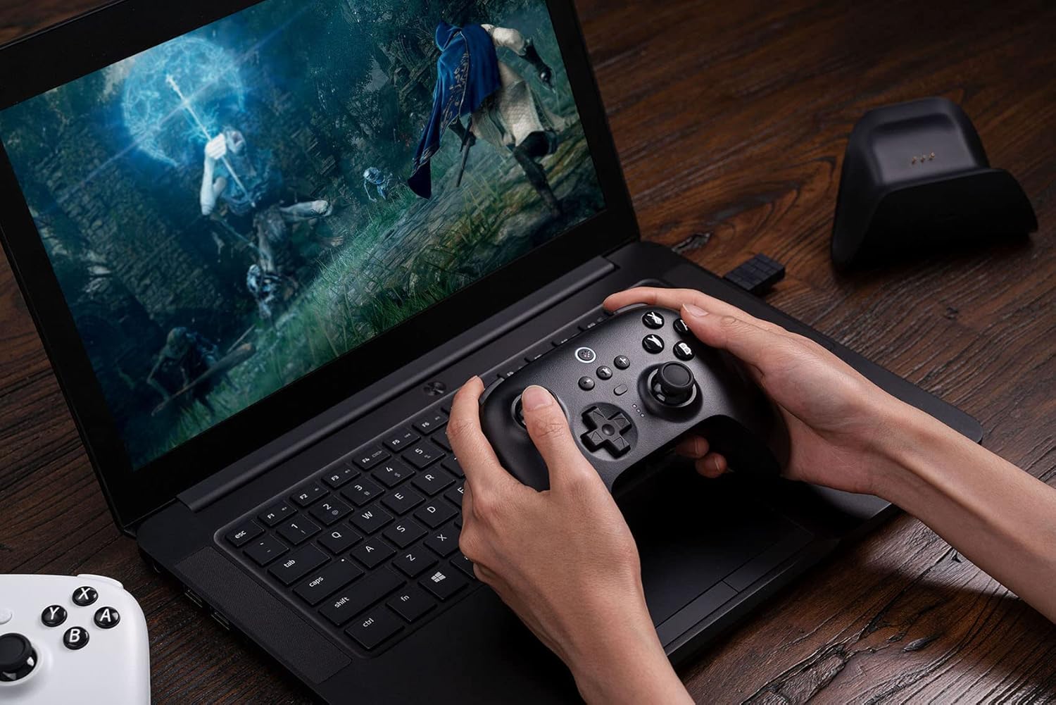 Hands holding an 8BitDo Ultimate Controller next to a laptop showing a game.