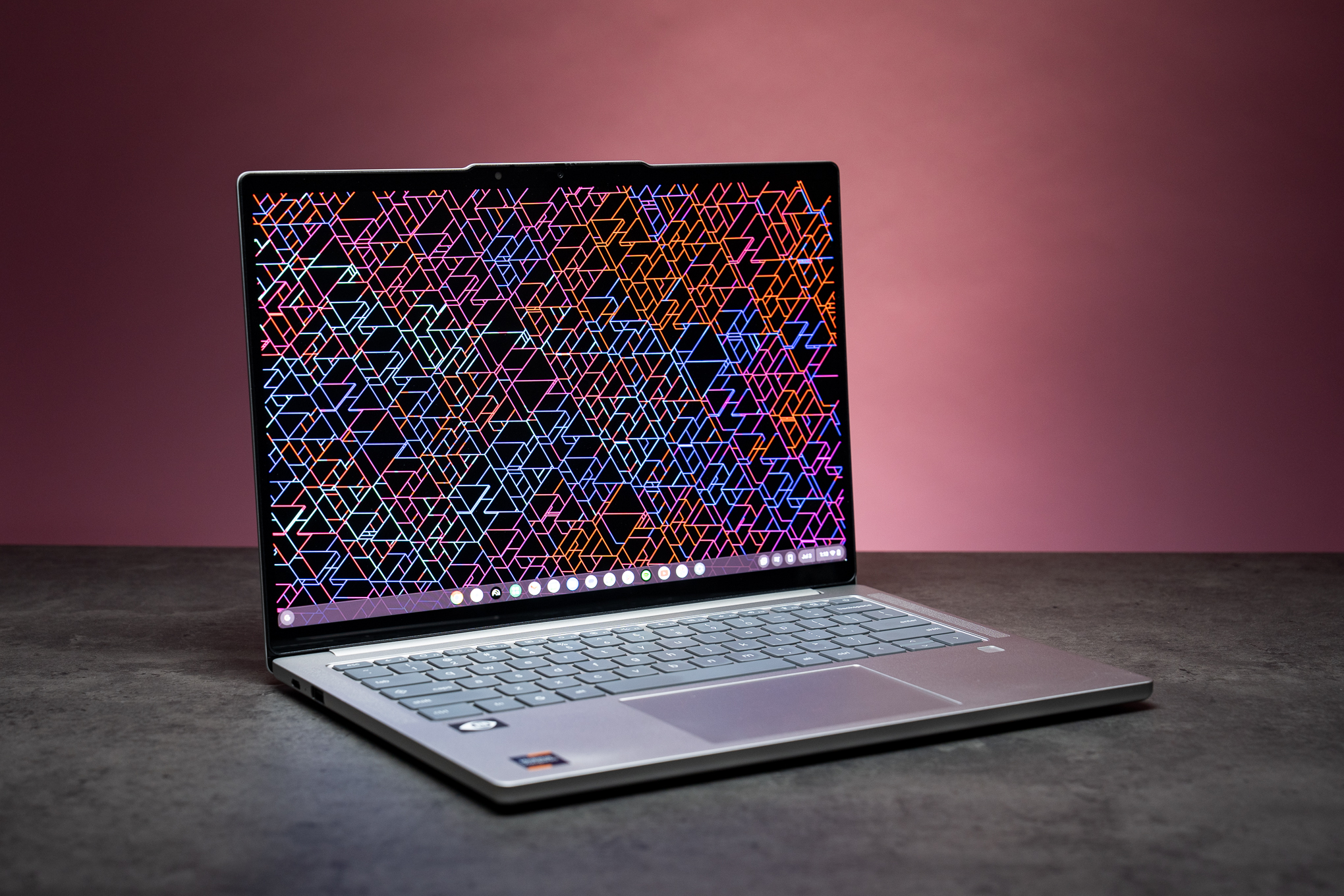 A Lenovo Chromebook Plus 14 laptop on a gray table in front of a pale pink background.