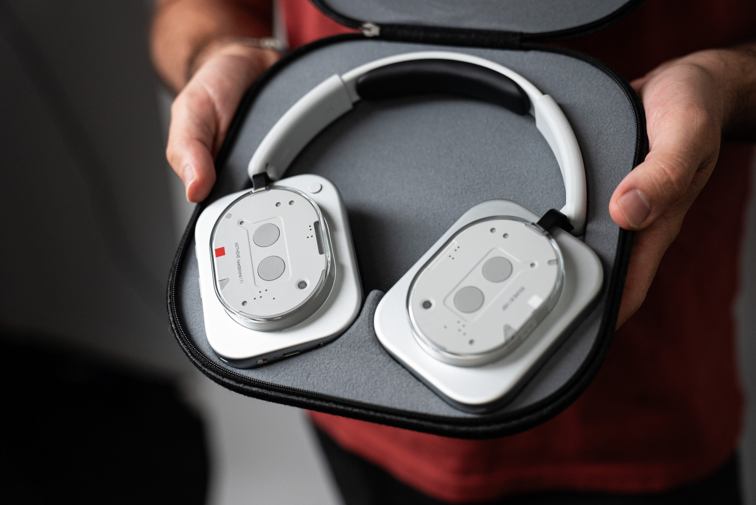 Two hands holding out the Nothing headphones, lying flat in the soft-shell case.