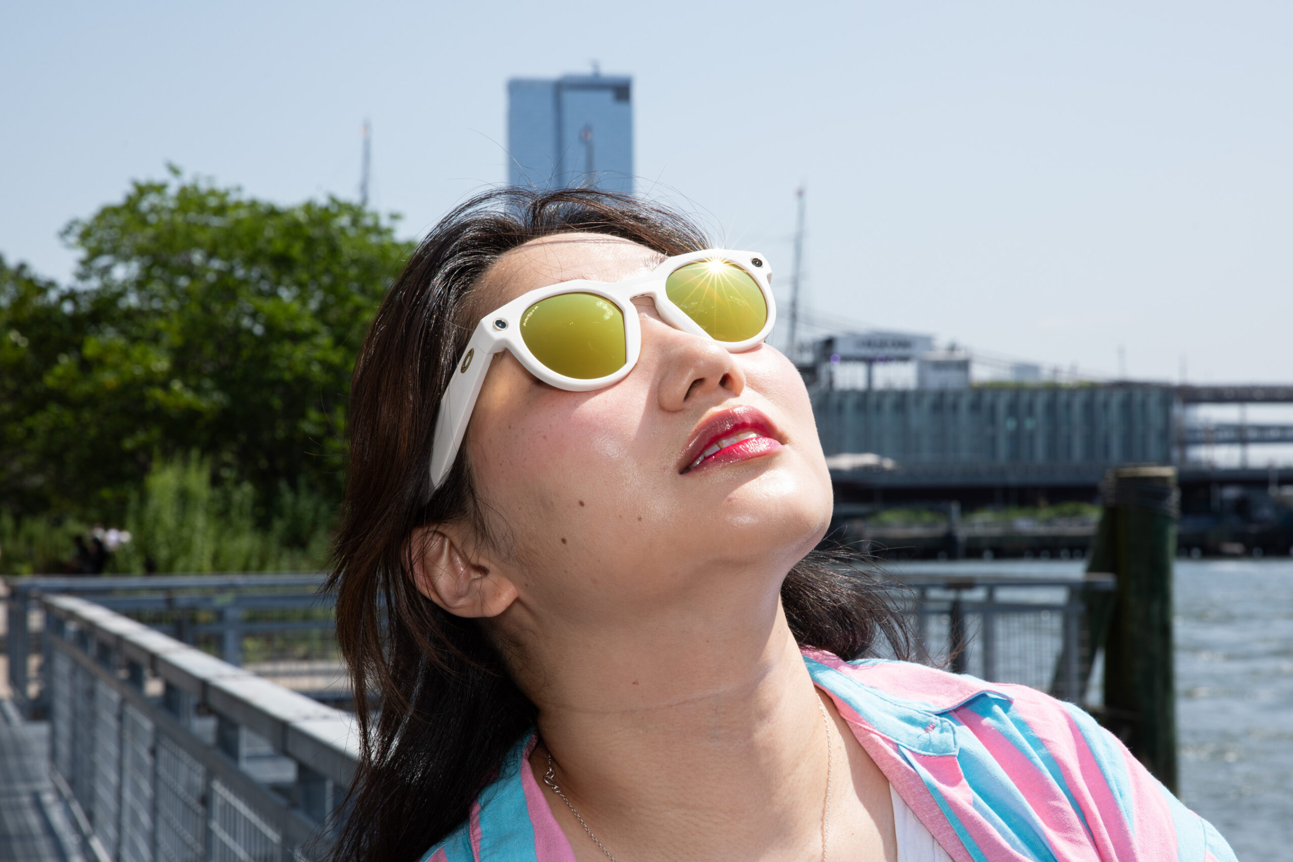 Senior reviewer Victoria Song wearing the Oakley Meta HSTN Limited Edition glasses while looking up at the sun. The glasses have a chunky white frame and polarized gold lenses. You can see a pier in the background.