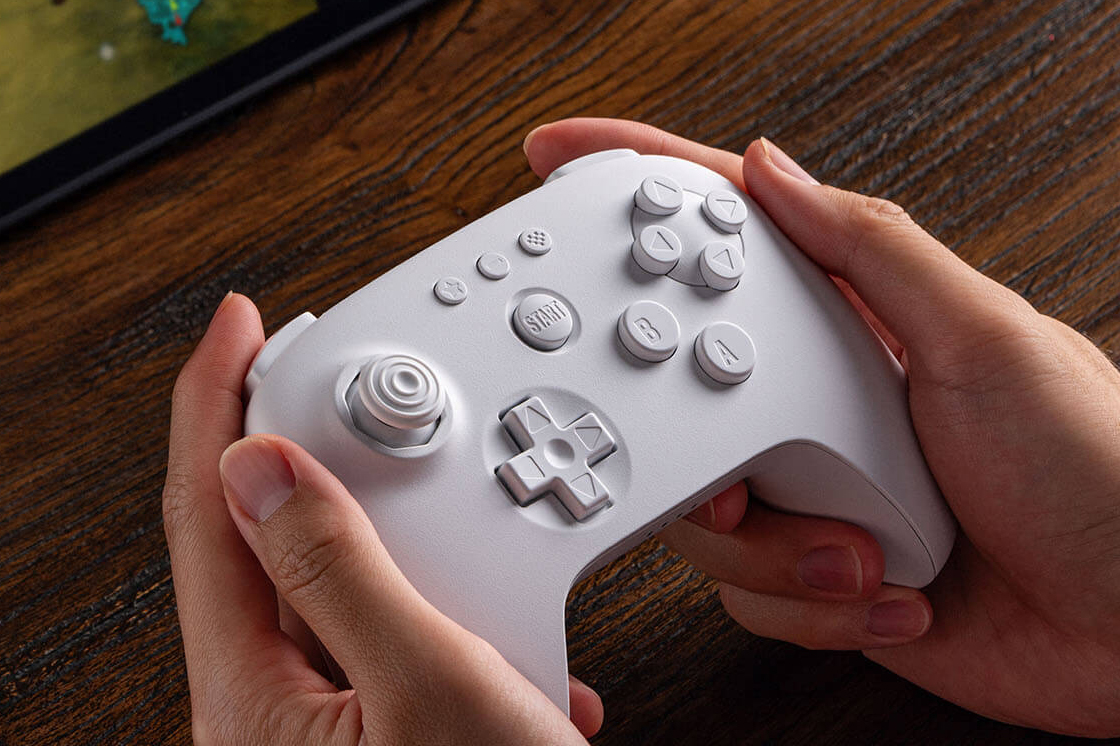 A hand holds the white version of the 8BitDo 64 controller.