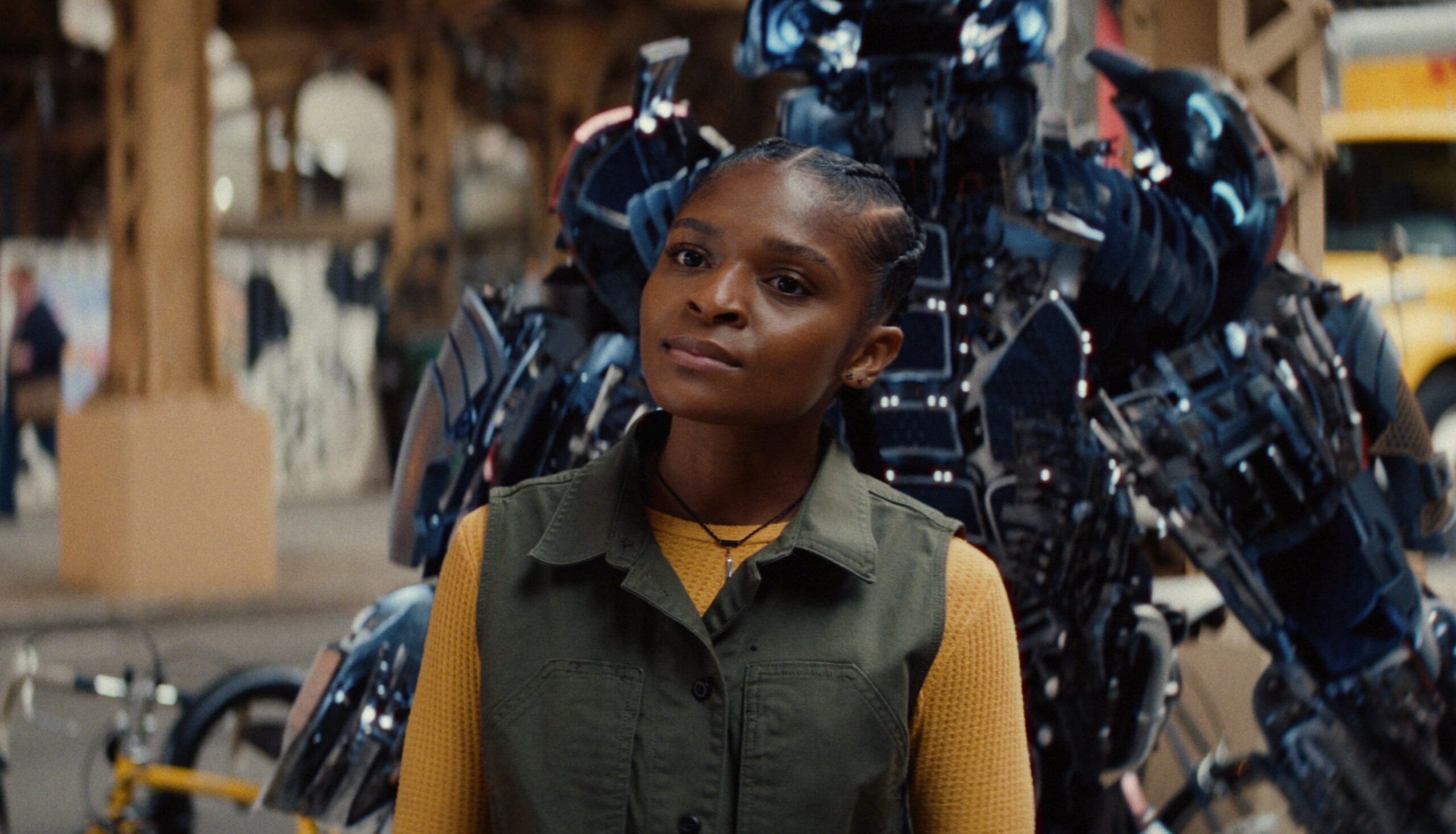 A girl in a yellow shirt and green vest standing on front of a human-shaped power armor that has opened up.