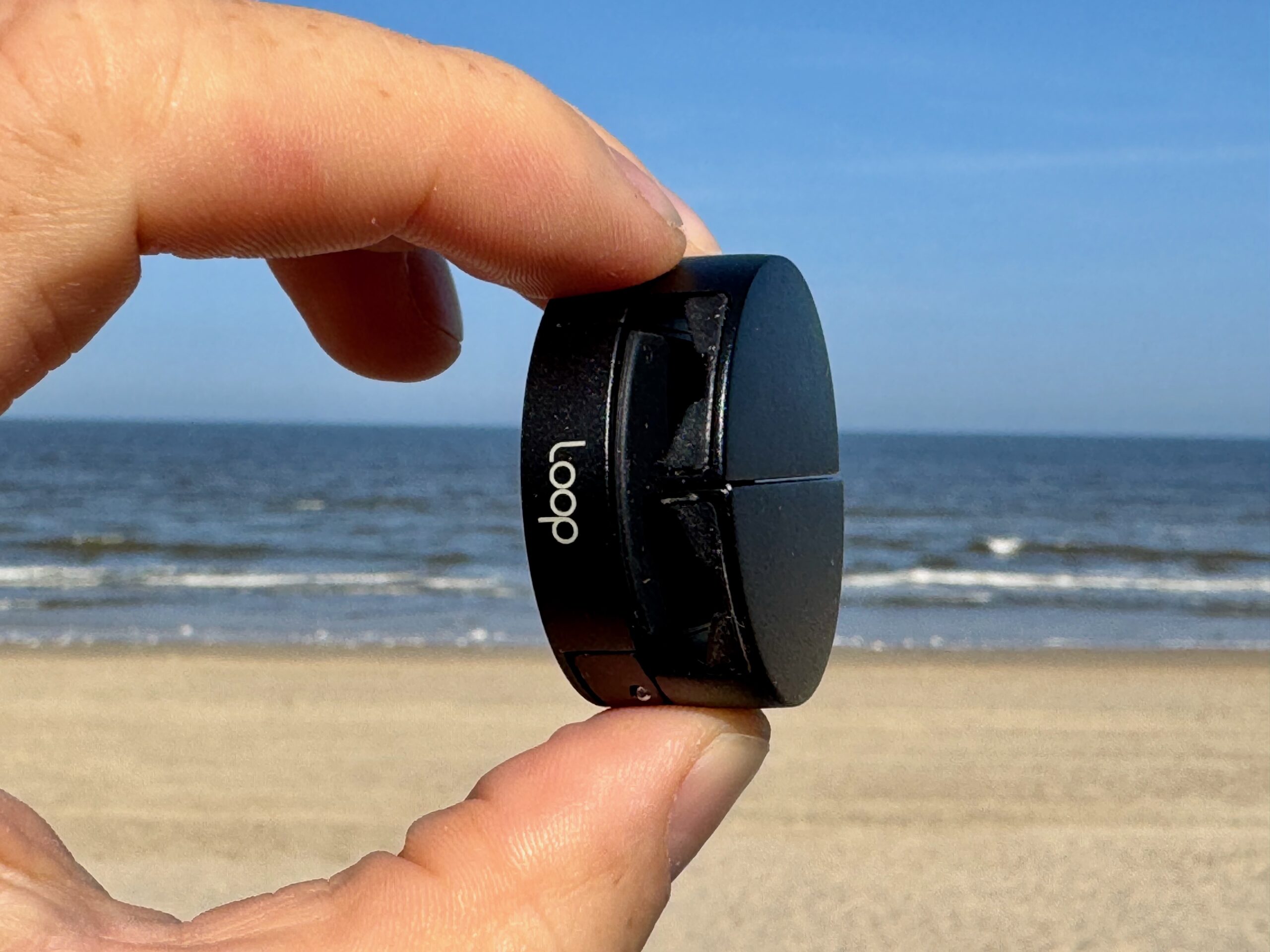 The circular Loop Micro Mount held between two fingers with the beach, ocean, and sky in the background.