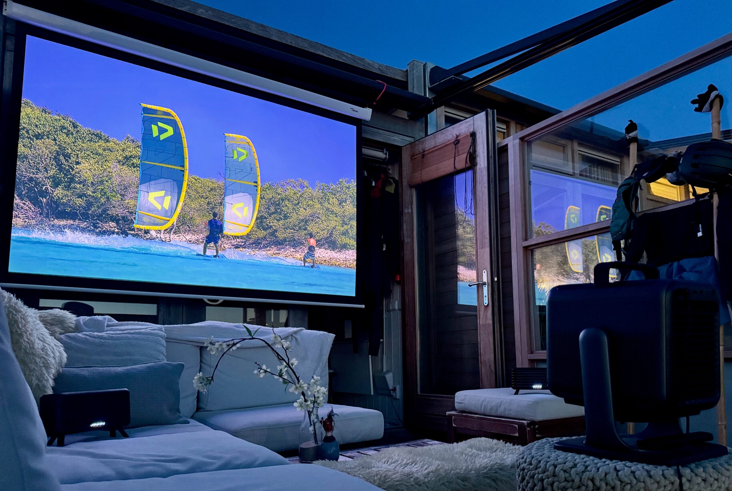 A low-angle shot captures an outdoor home theater at dusk. A large projection screen on the left shows two people kitesurfing on turquoise water next to a lush green coastline, with two bright yellow and blue kites prominently displayed. In the foreground, a comfortable outdoor sofa with white cushions and throws is visible, along with a white ottoman. To the right, the Nebula X1 projector sits on a textured, light-colored stool. Wooden structures and glass panels enclose the space, reflecting the evening light. A small white floral arrangement is on a table between the sofa and the projector.