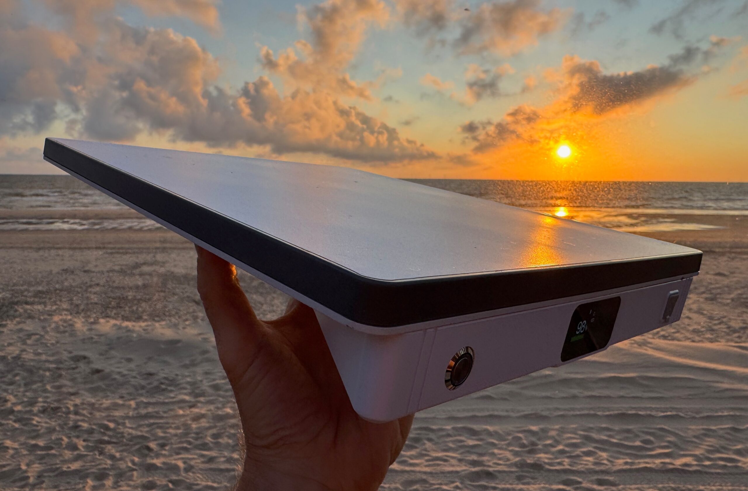 The PeakDo LinkPower inserted into the back of the Starlink Mini and the combined unit held aloft by a hand at sunset with the ocean, beach, and dramatic sky visible in the background.