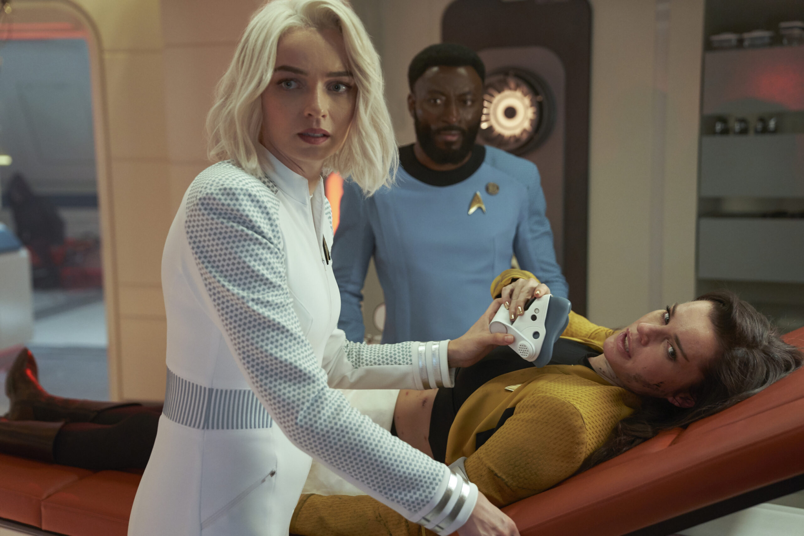 A dark haired White woman lies on a stretcher being checked for injured by a blond White woman, and a dark haired Black man looks on with concern.