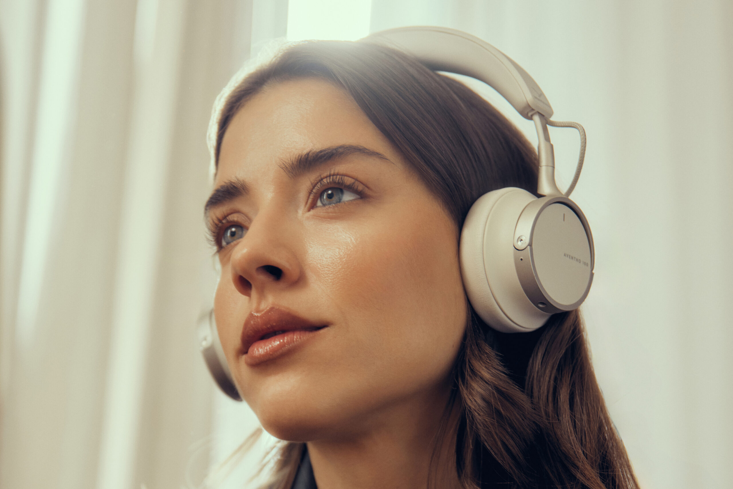 A person wears the cream version of the Beyerdynamic Aventho 100 wireless headphones in front of a hanging curtain.