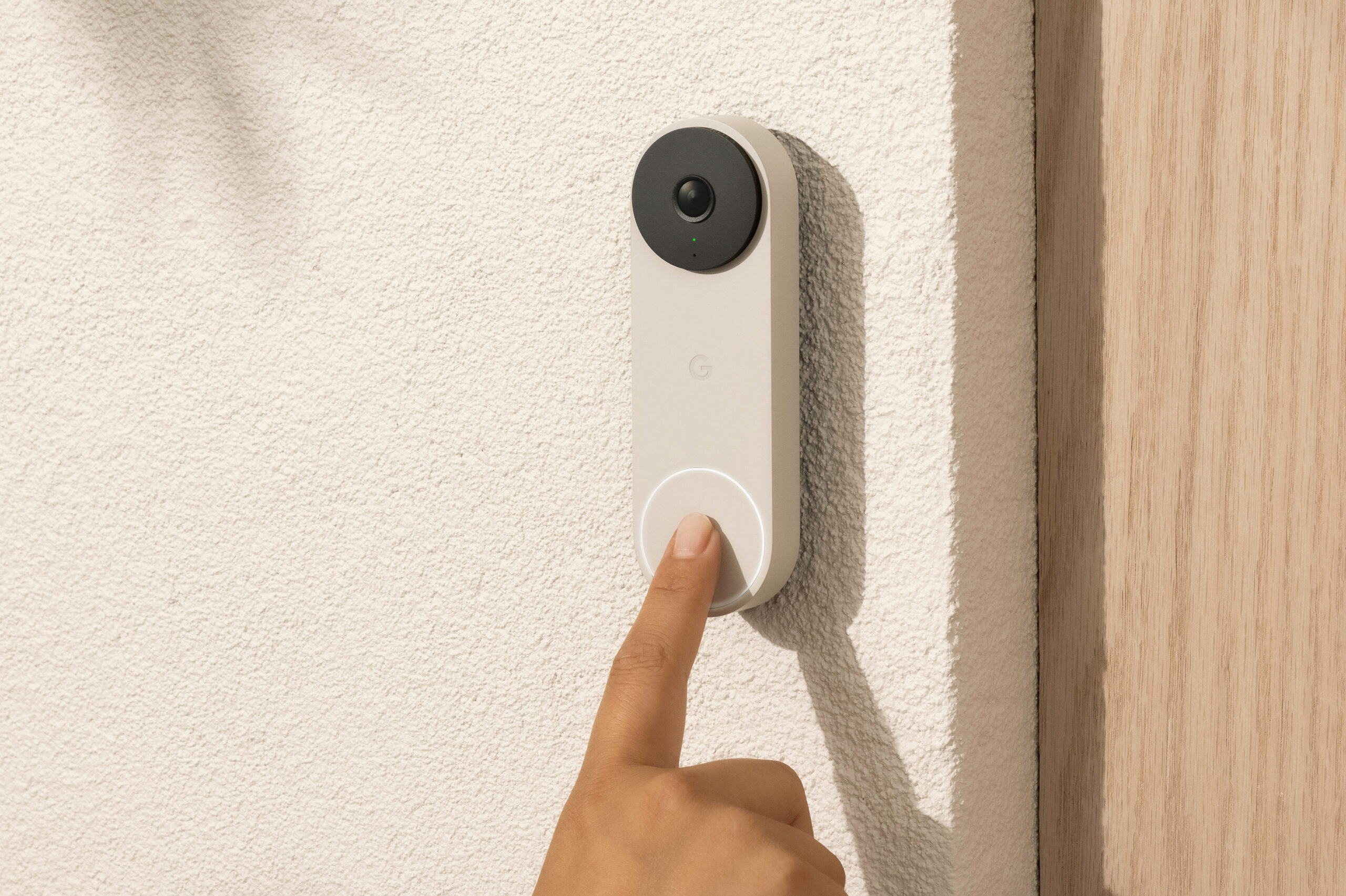 A finger presses a button on a Nest Doorbell attached to a wall next to a door.