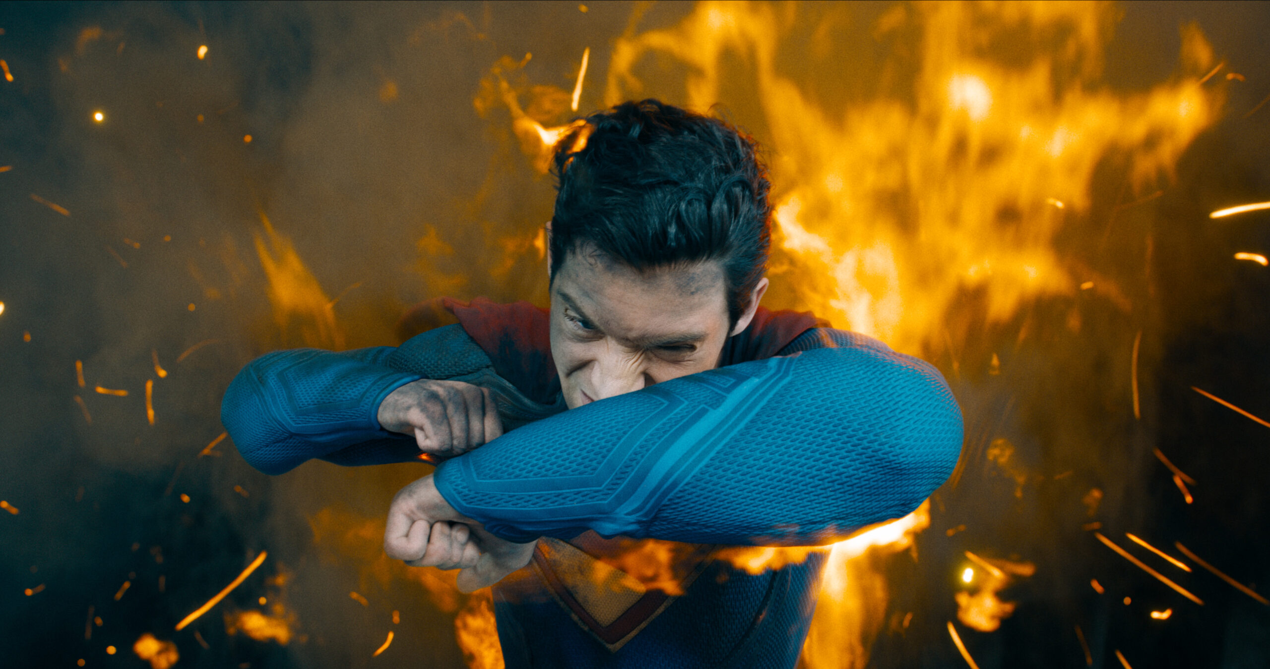 Superman in his classic blue and red suit shielding his face from debris as something explodes behind him.