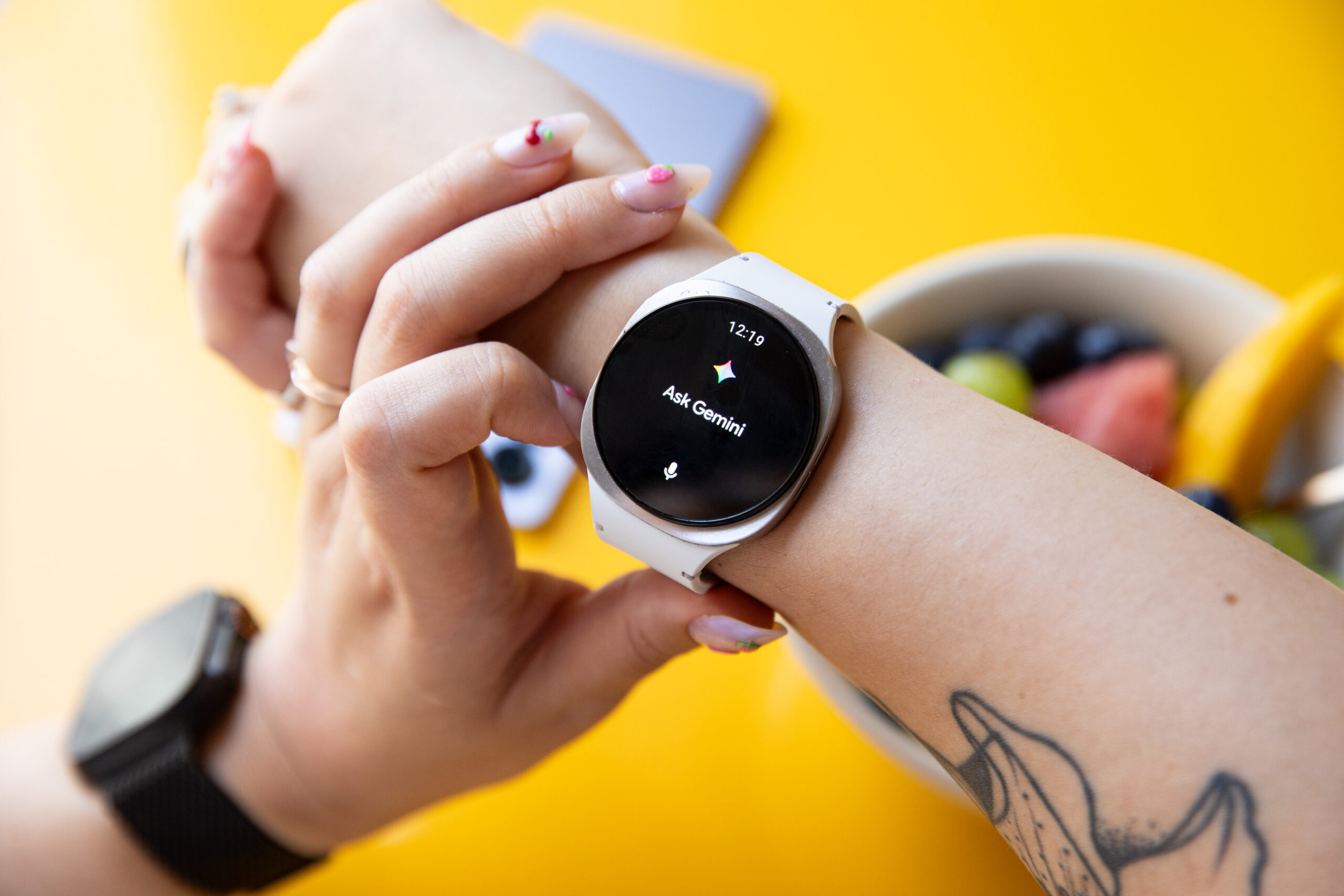 Close up of the Samsung Galaxy Watch 8 on Senior Reviewer Victoria Song’s wrist showing the “Ask Gemini” screen. There’s a yellow table and a bowl of fruit in the background. 