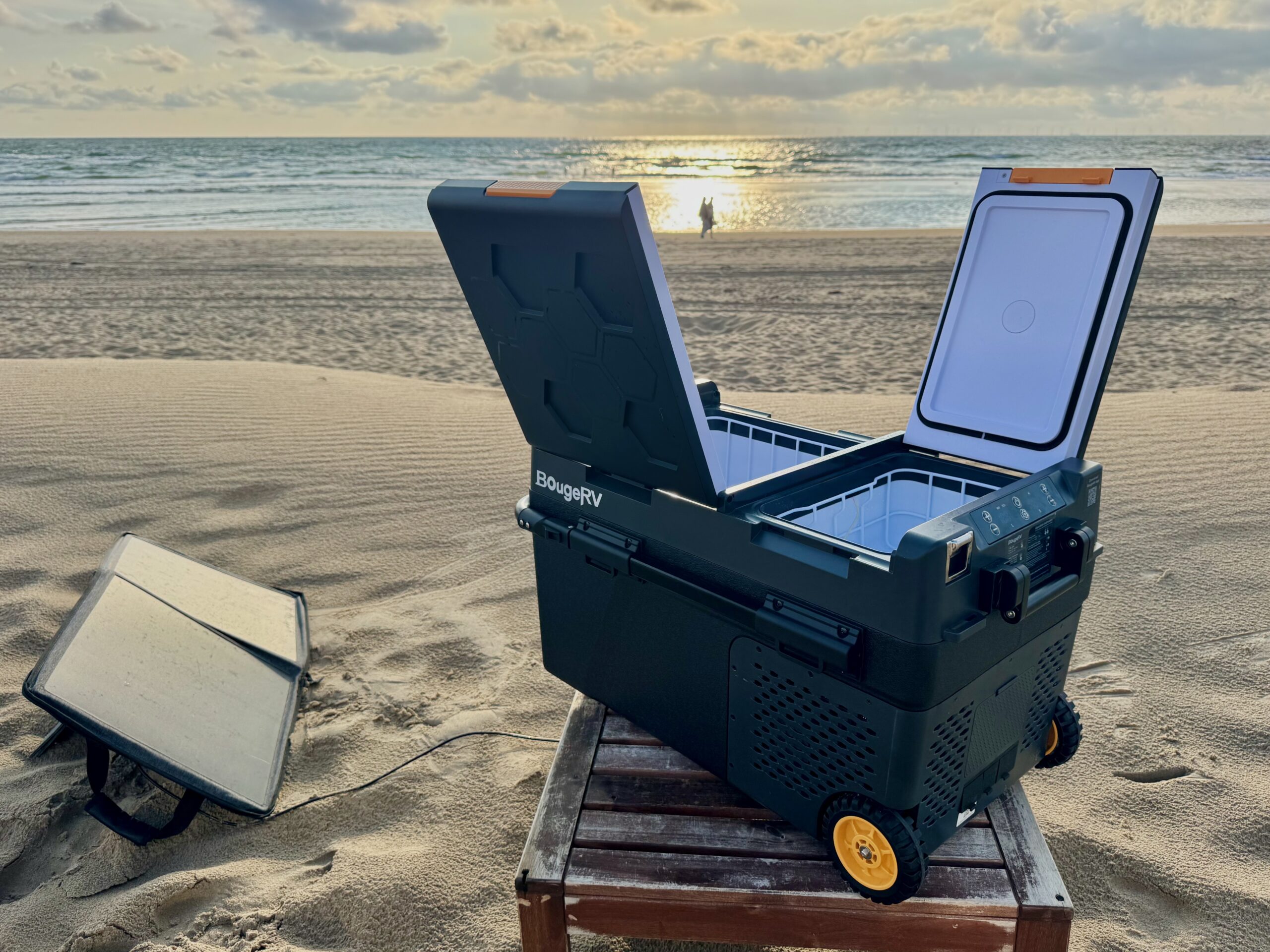 The BougeRV fridge sitting on a short table on the beach with a solar panel nearby as the sun reflects off the ocean in the distance.