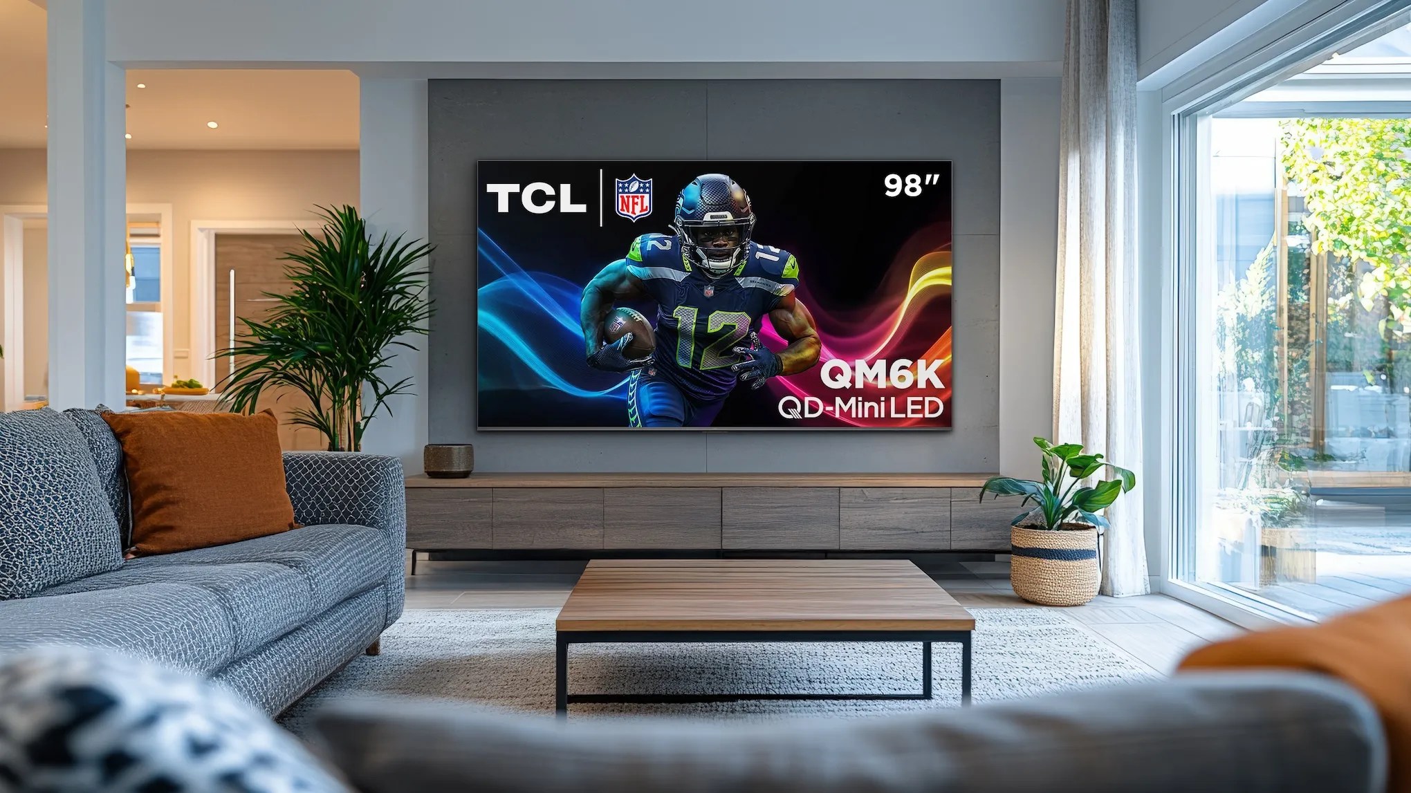 An image of the 98-inch TCL QM6K QD-Mini LED TV mounted on the wall in a cozy living room with a lot of natural light coming from nearby windows.
