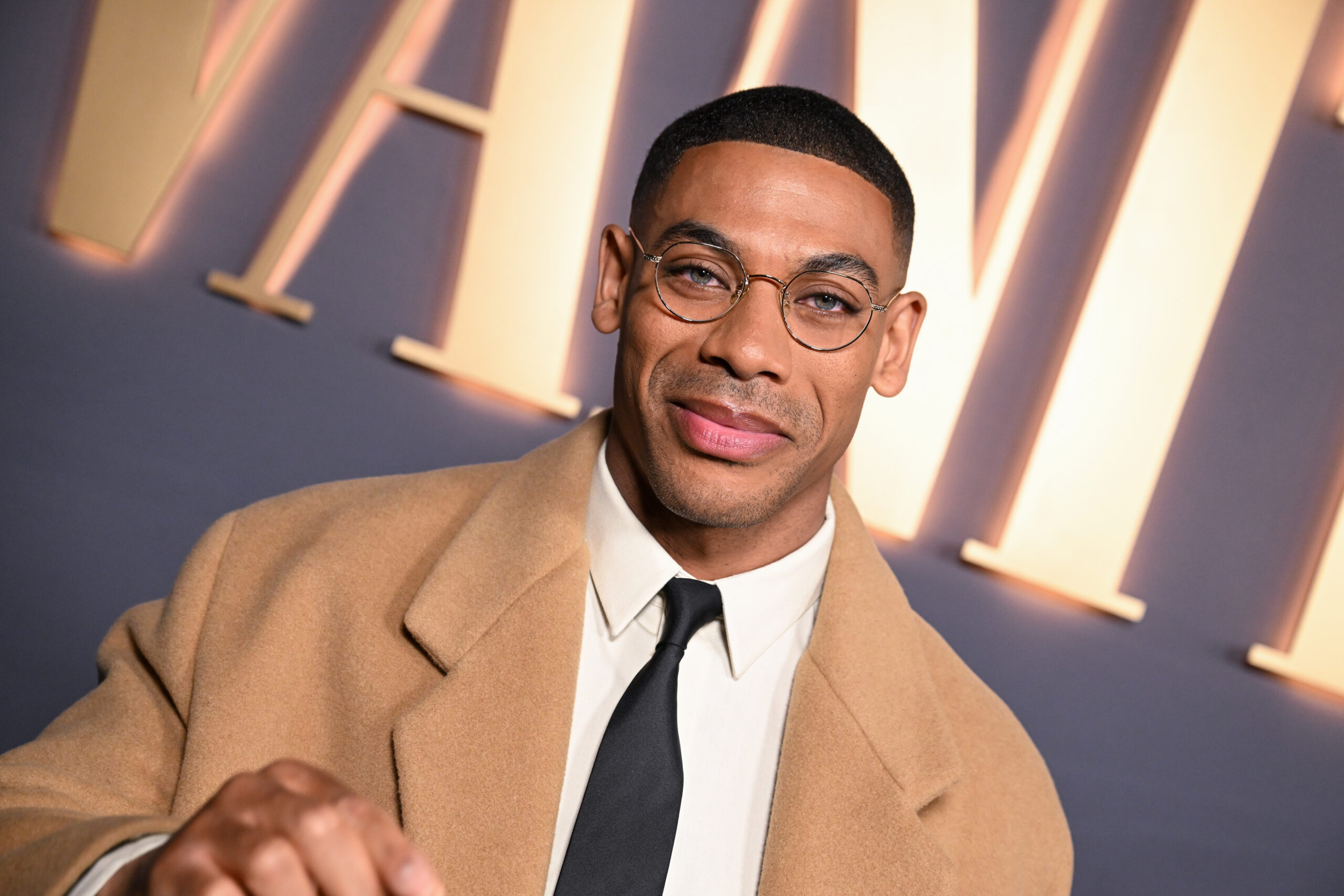 Actor Aaron Pierre in a tan suit standing on a red carpet.