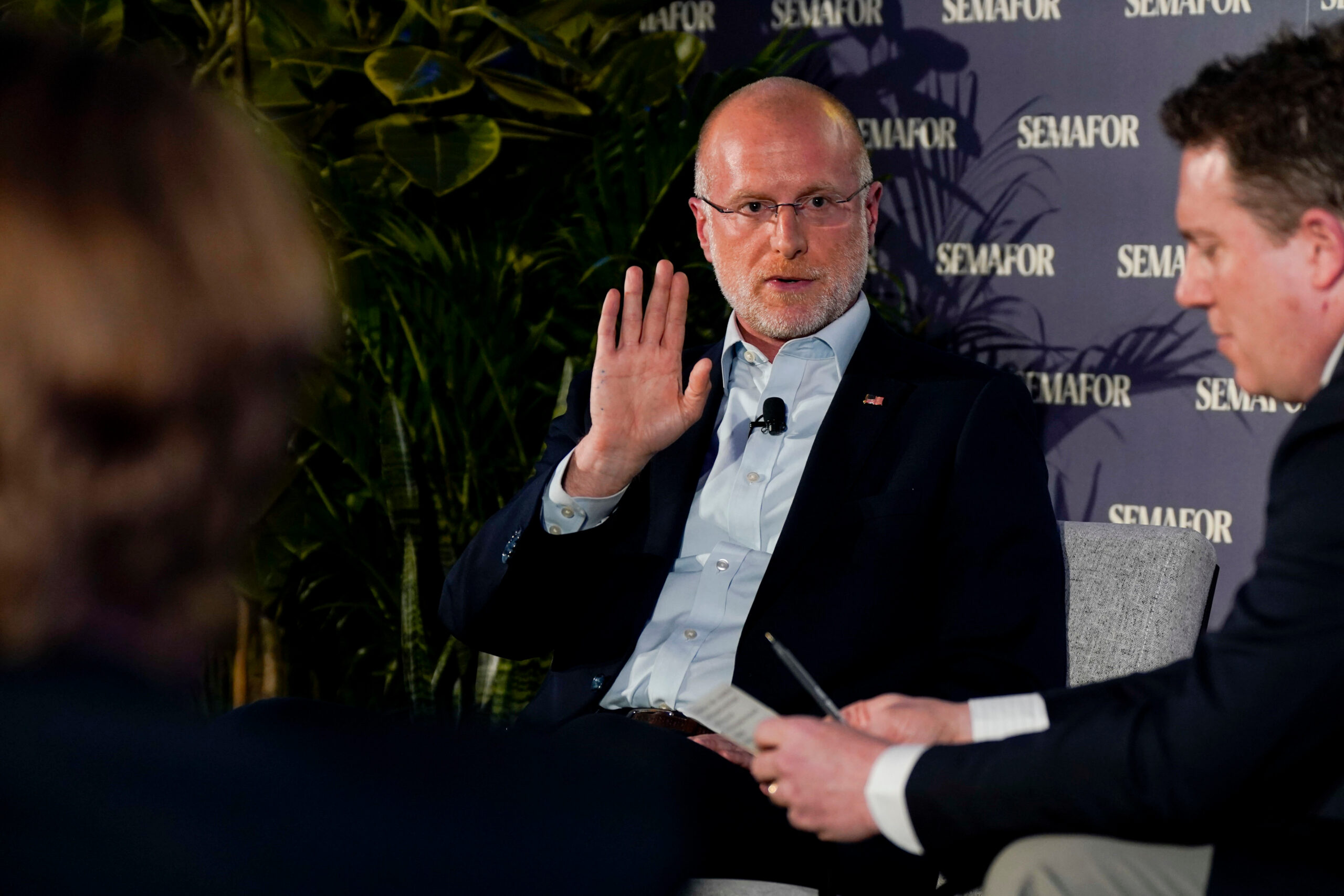 Brendan Carr, FCC chairman, raises his hand in a “stop”-like gesture while sitting in a chair.