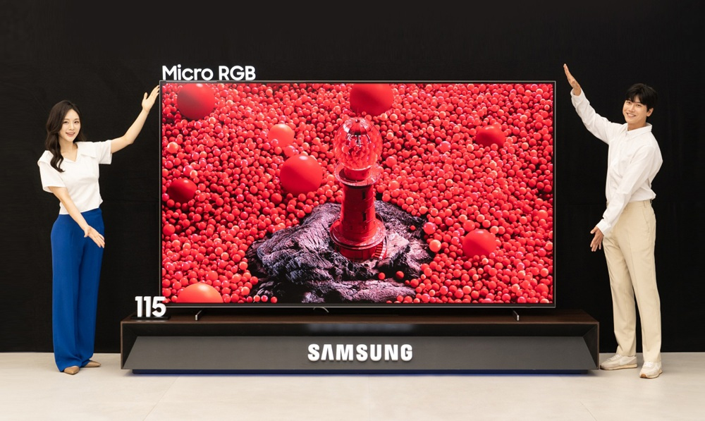 Two people stand on either side of a 115-inch Samsung Micro RGB TV showing red bubbles on screen.