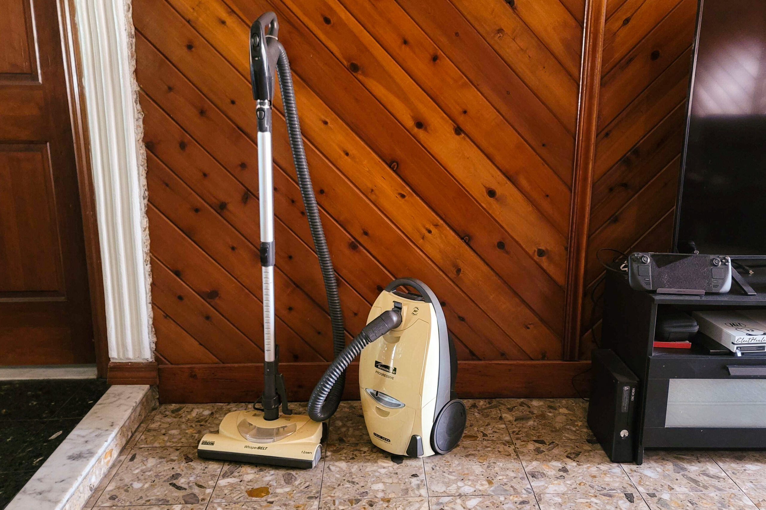 Old-fashioned vacuum cleaner with a canister next to it on the floor with a wood-paneled wall behind it.