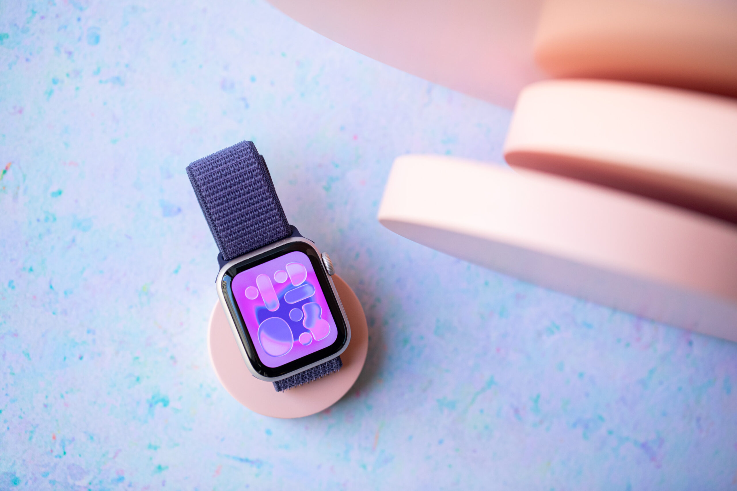 Apple Watch SE 3 on a peach colored disc with more discs standing vertically to the right side. There’s a speckled blue background.