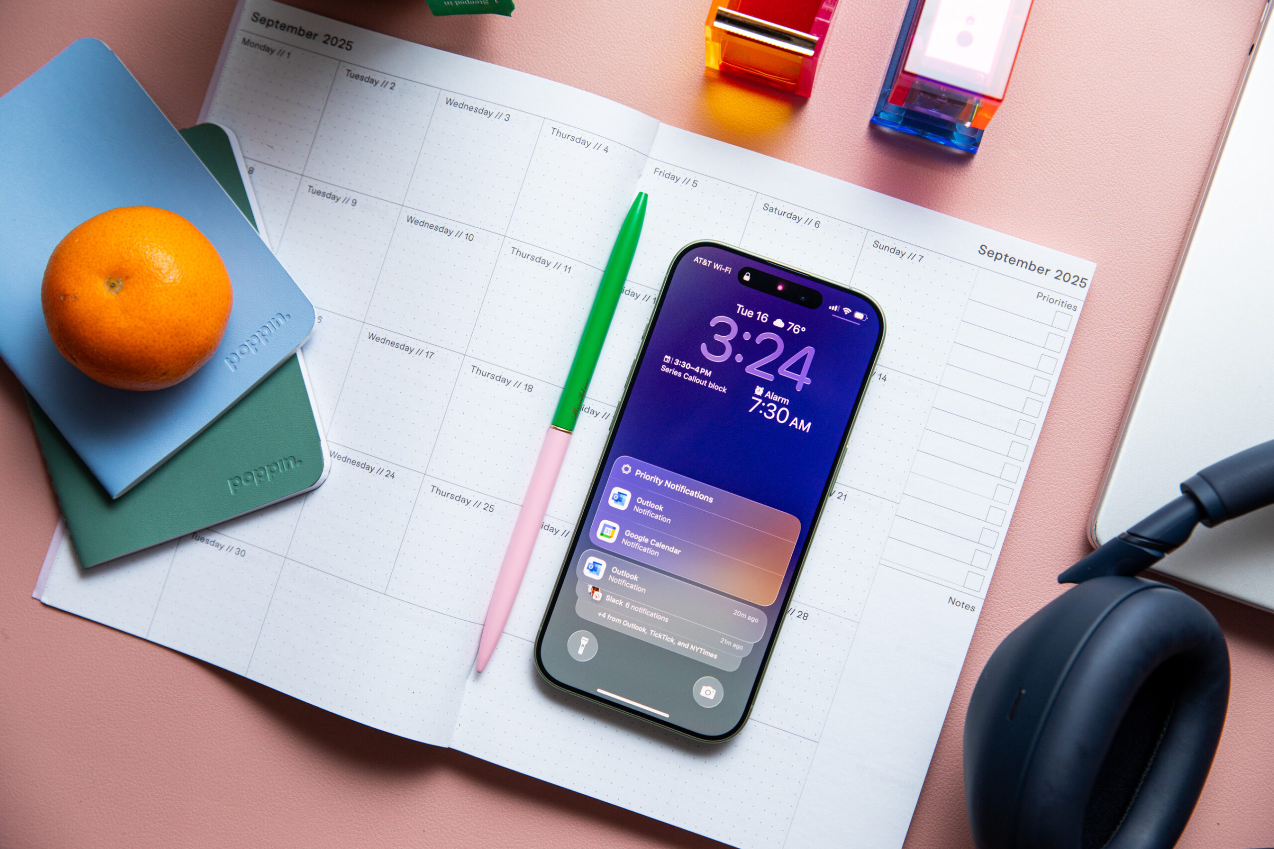 An iPhone resting on a blank monthly planner, next to a pink and green pencil. Several notebooks and an orange are in the shot. It’s nice. 