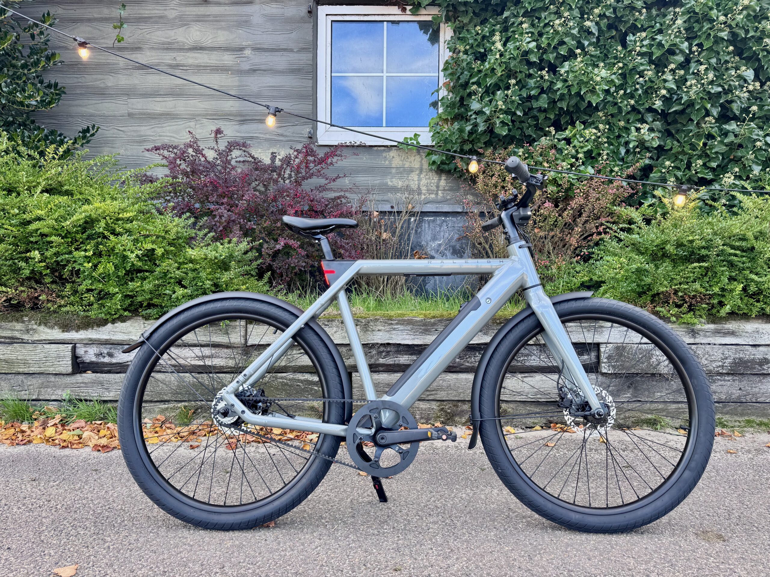 The Raleigh One e-bike in front of wooden house and window dotted lined with foilage and a string of outdoor lights.