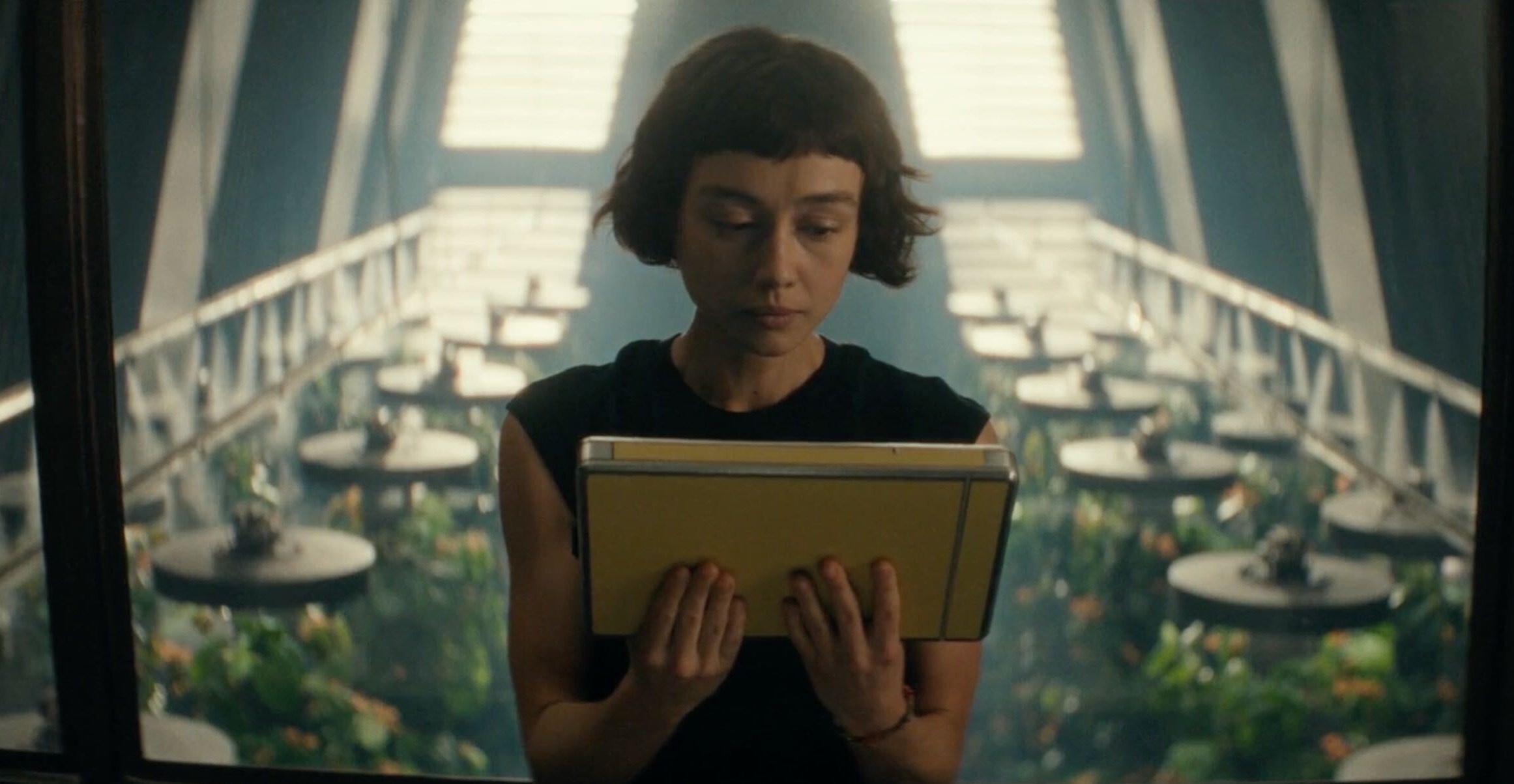 A woman sitting in a room above an indoor garden. The woman is using both hands to hold a chunky tablet that she is looking at.