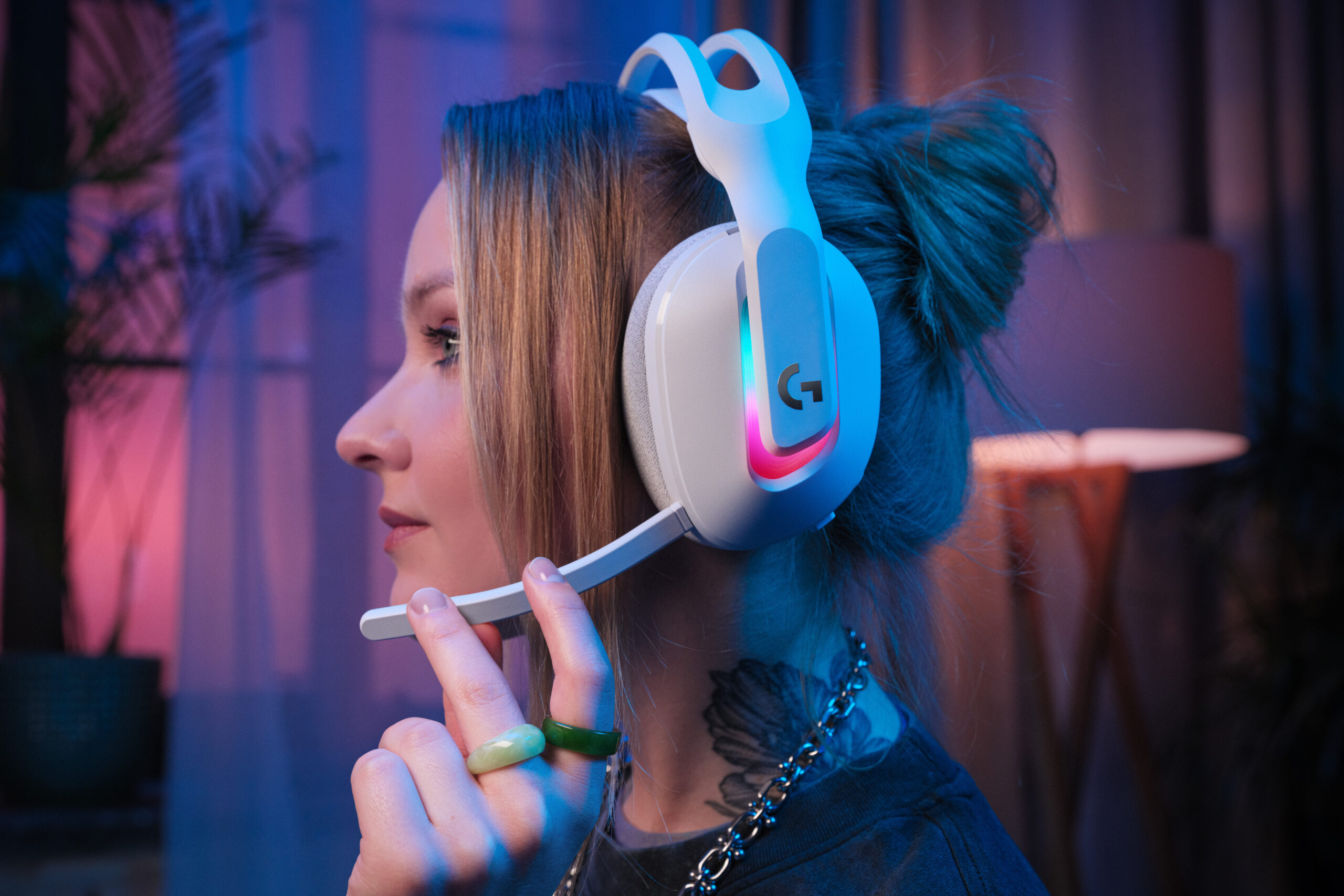 An image showing a model wearing the Astro A20 X gaming headset while turned to the side. They are in a dimly lit room, and pink and purples accents are emphasized throughout.