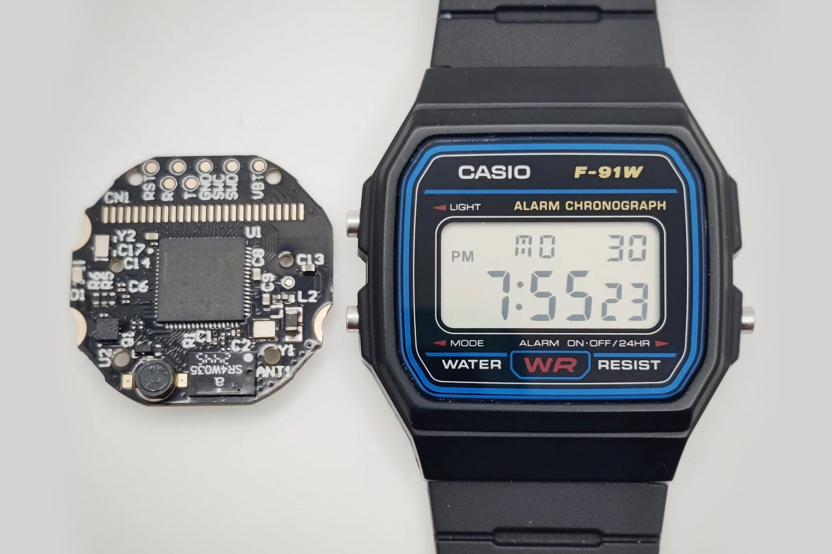 A Casio F-91W digital watch next to the Ollee Watch One replacement mainboard.