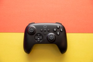 Top-down shot of 8BitDo Ultimate Controller on an orange and yellow background. The controller has an asymmetric joystick design, like an Xbox or Switch Pro controller, and it’s black, with white legends on the A B X Y buttons.