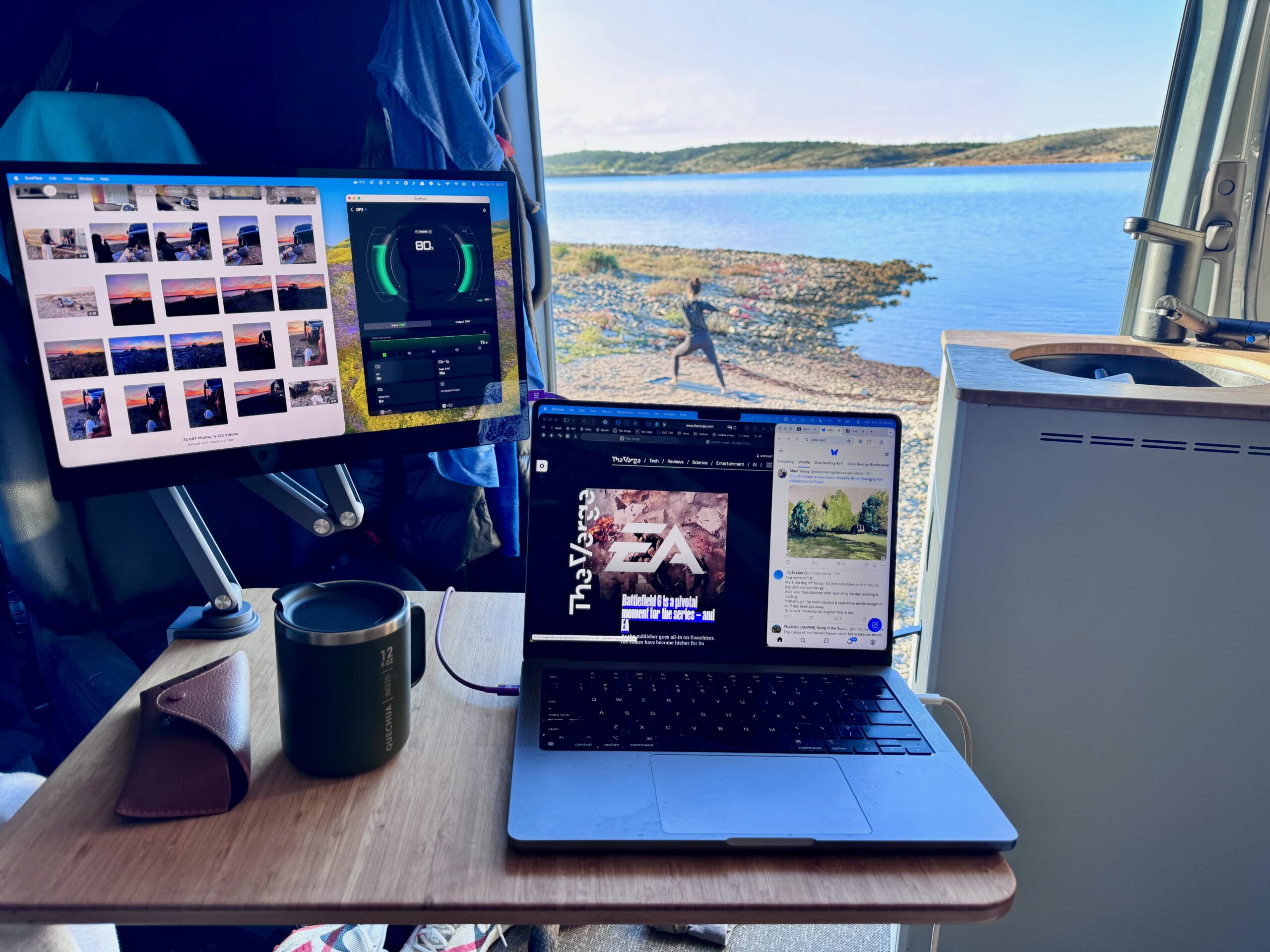 The Espresso Pro monitor, mounted onto a swiveling arm inside van, hovers above and to the left of the MacBook Pro placed on table next to a coffee cup and glasses. A woman does yoga next to the lake outside.