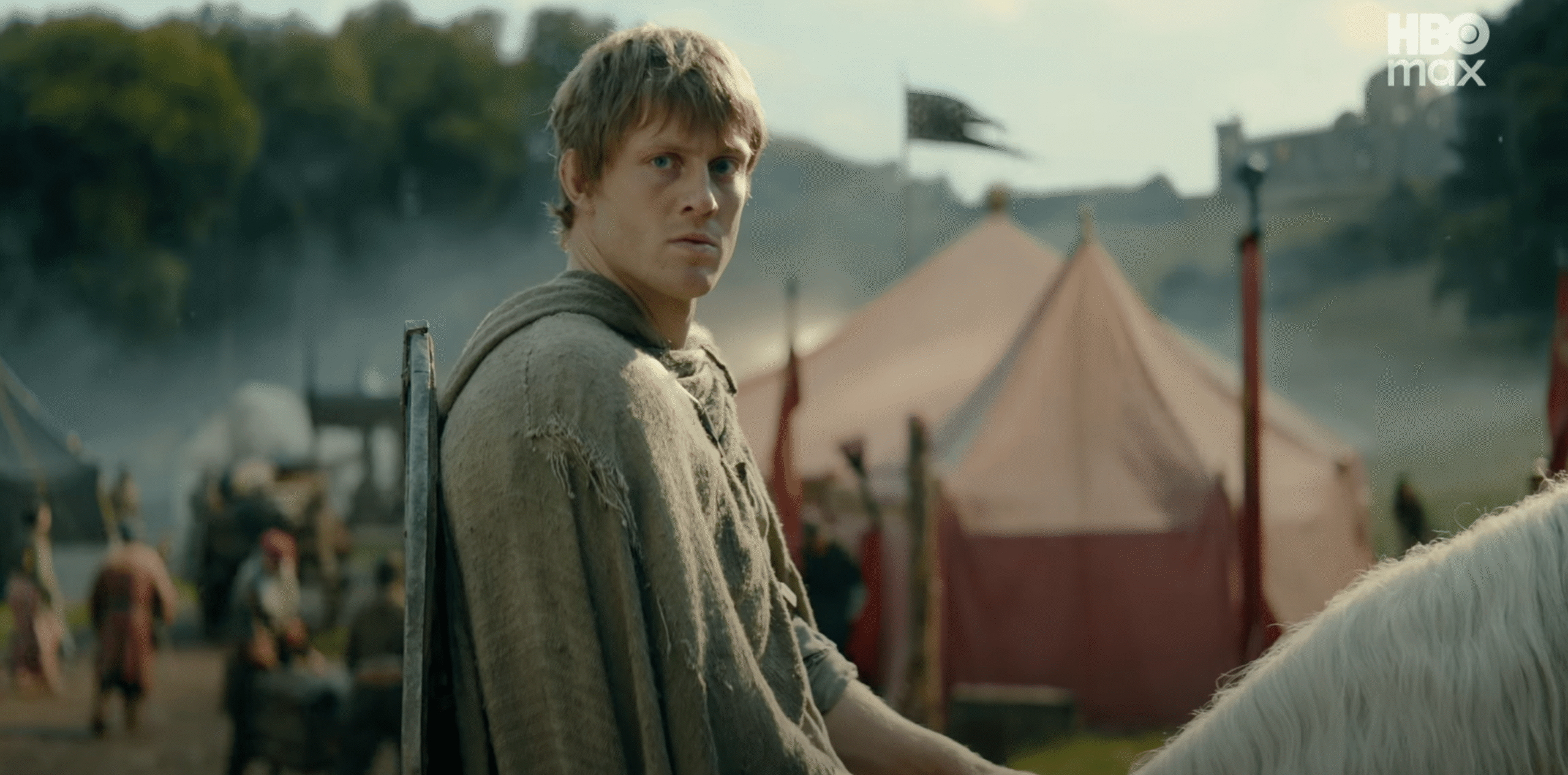 A man in simple knights clothing who is sitting on the back of a white horse. Behind the man is a red tent.