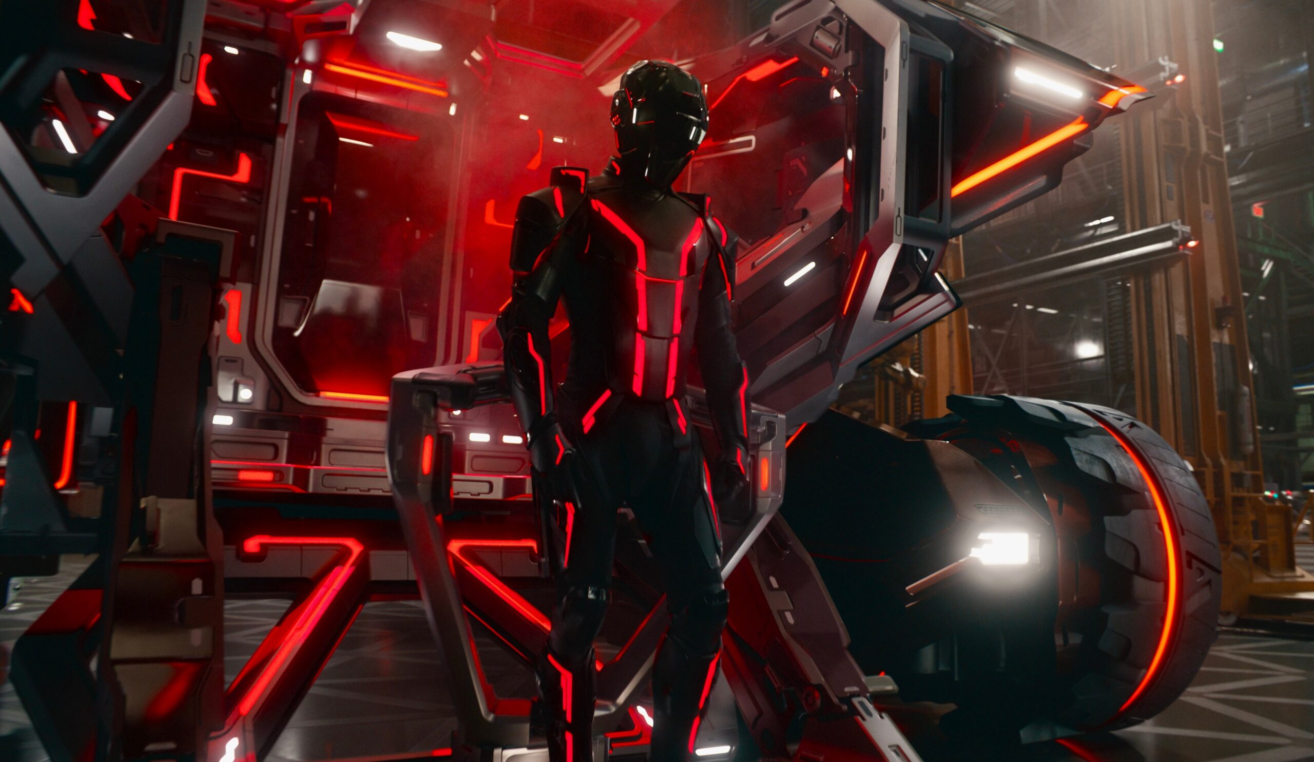 A man in form-fitting full body armor. The man is wearing a plastic mask and standing in the center of a futuristic tank accented with red light strips.